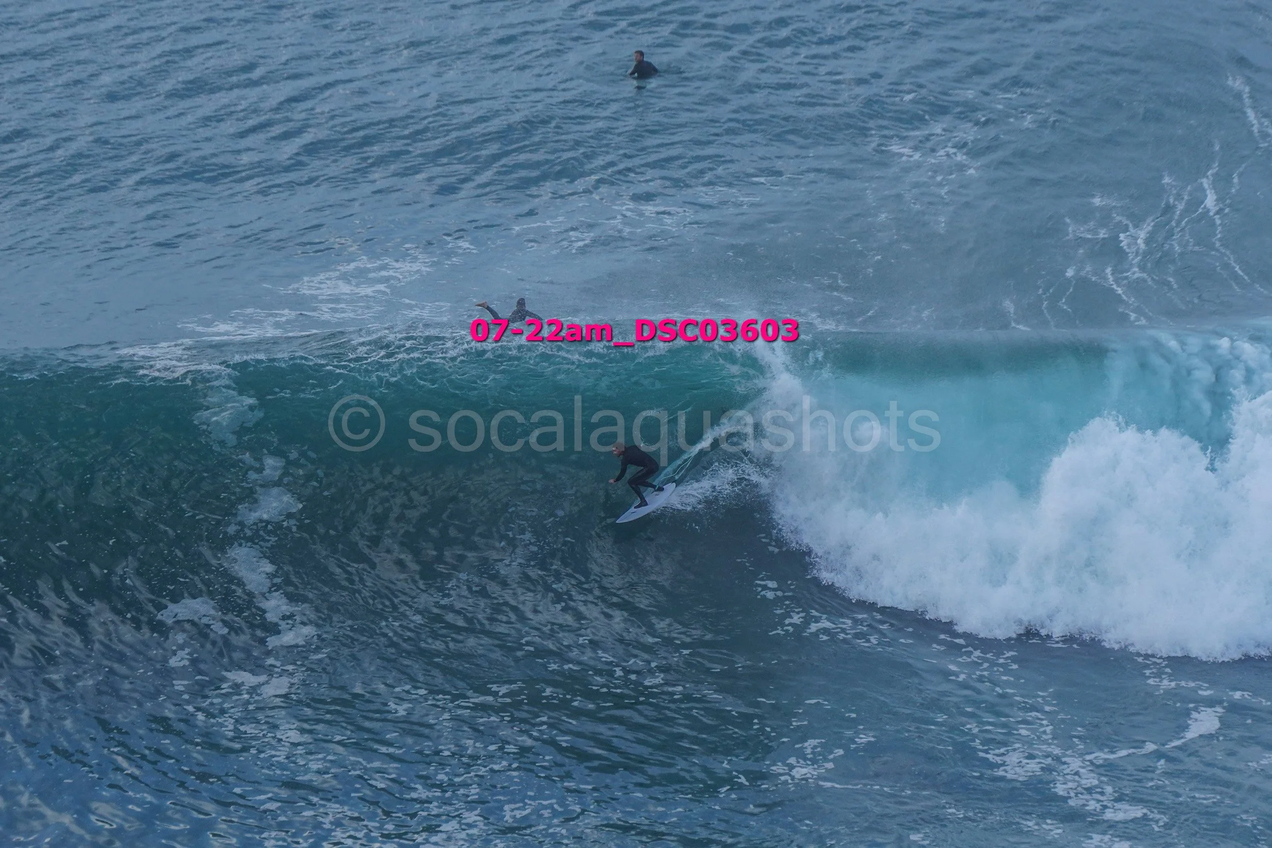 Surfer riding a large wave with three additional surfers visible in the water in the background.