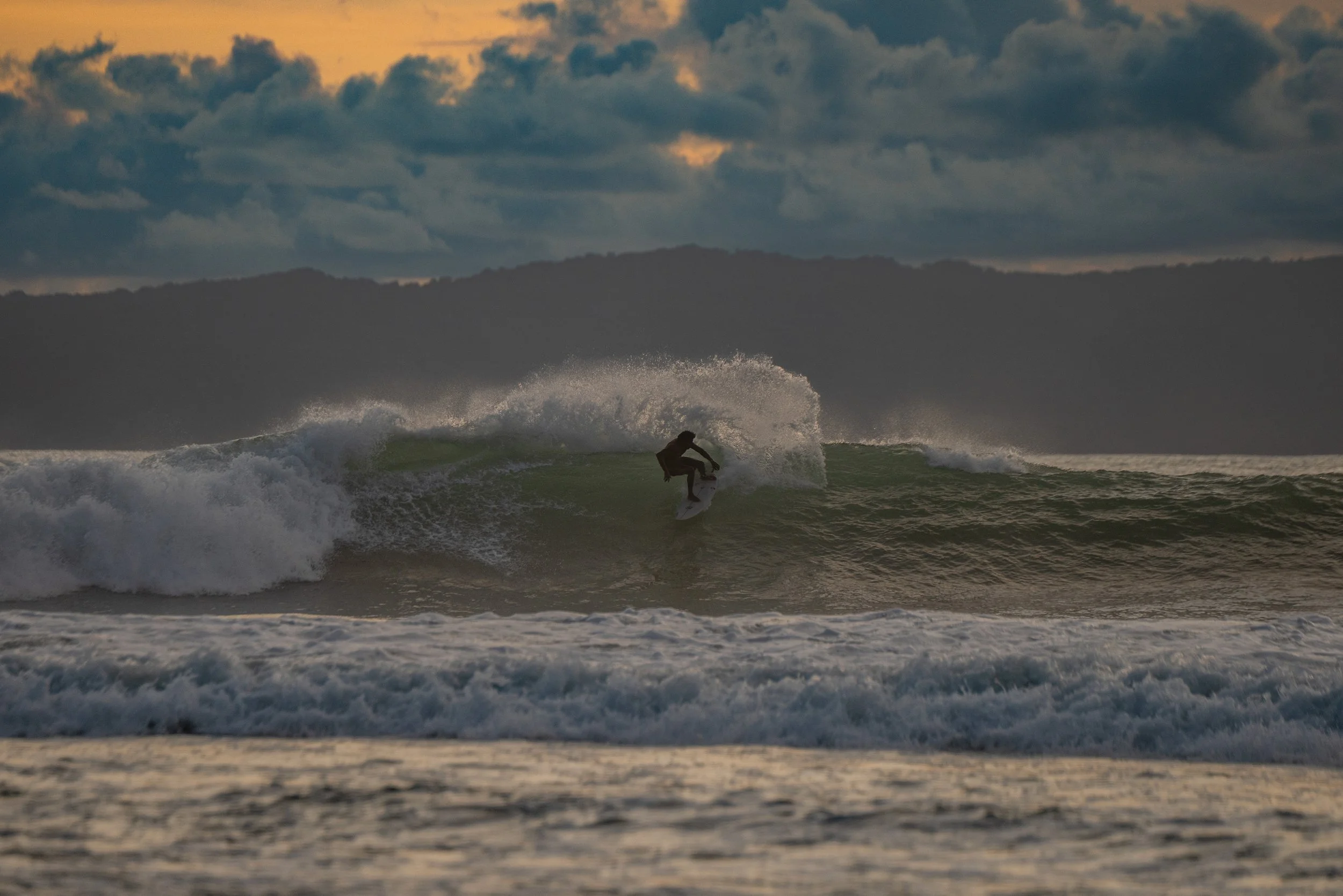 A person surfing on a wave in the ocean during sunset with dark clouds in the sky.