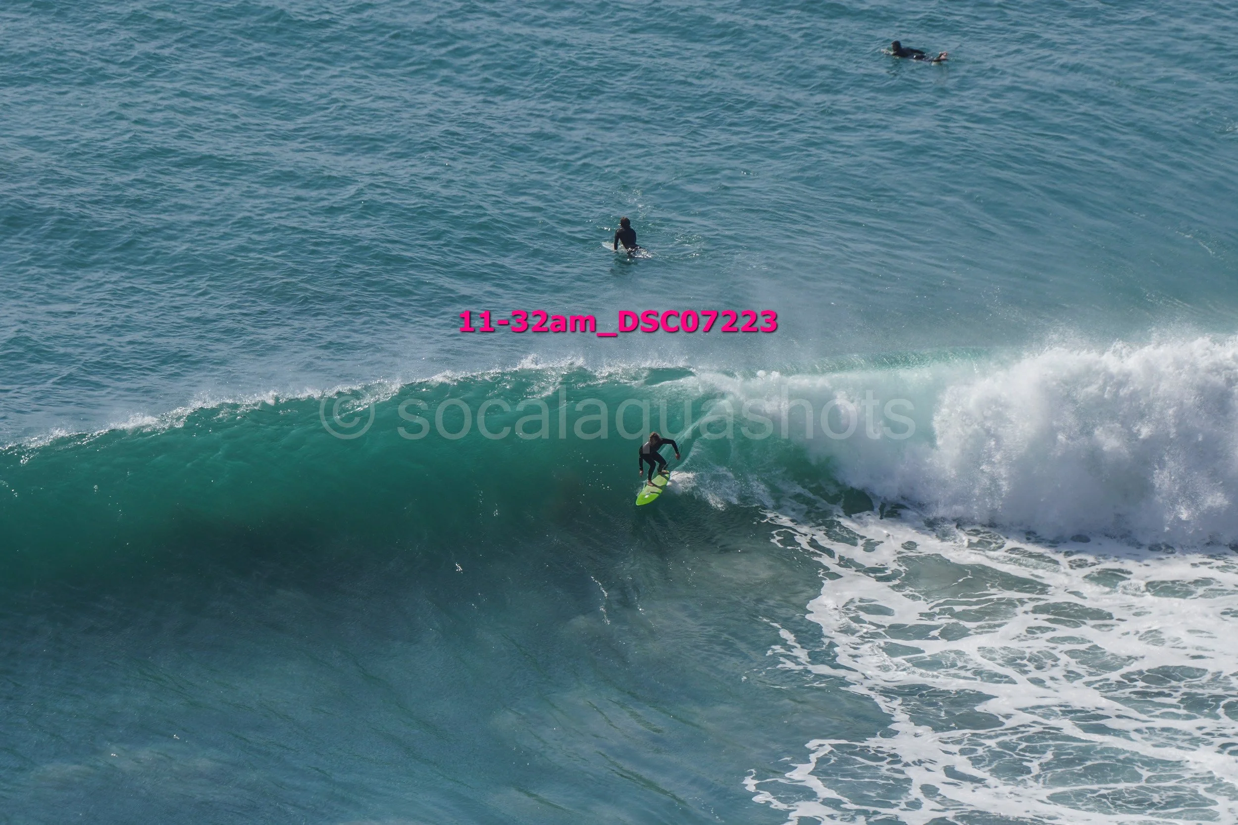A person surfing on a large wave with surfboard, two other individuals are in the water, one near the wave and one further out in the ocean.