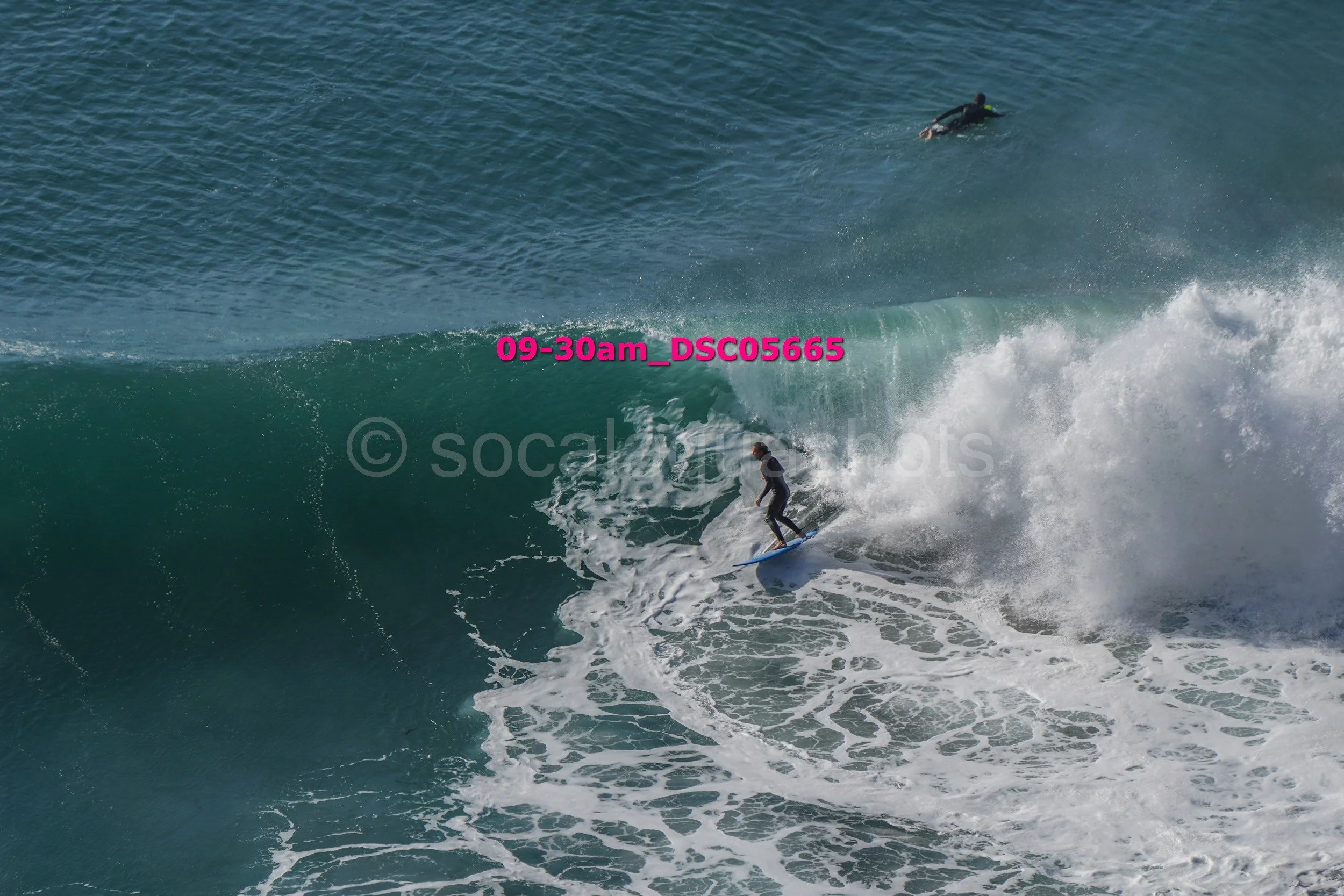 A person surfing on a large ocean wave with another swimmer in the water nearby.