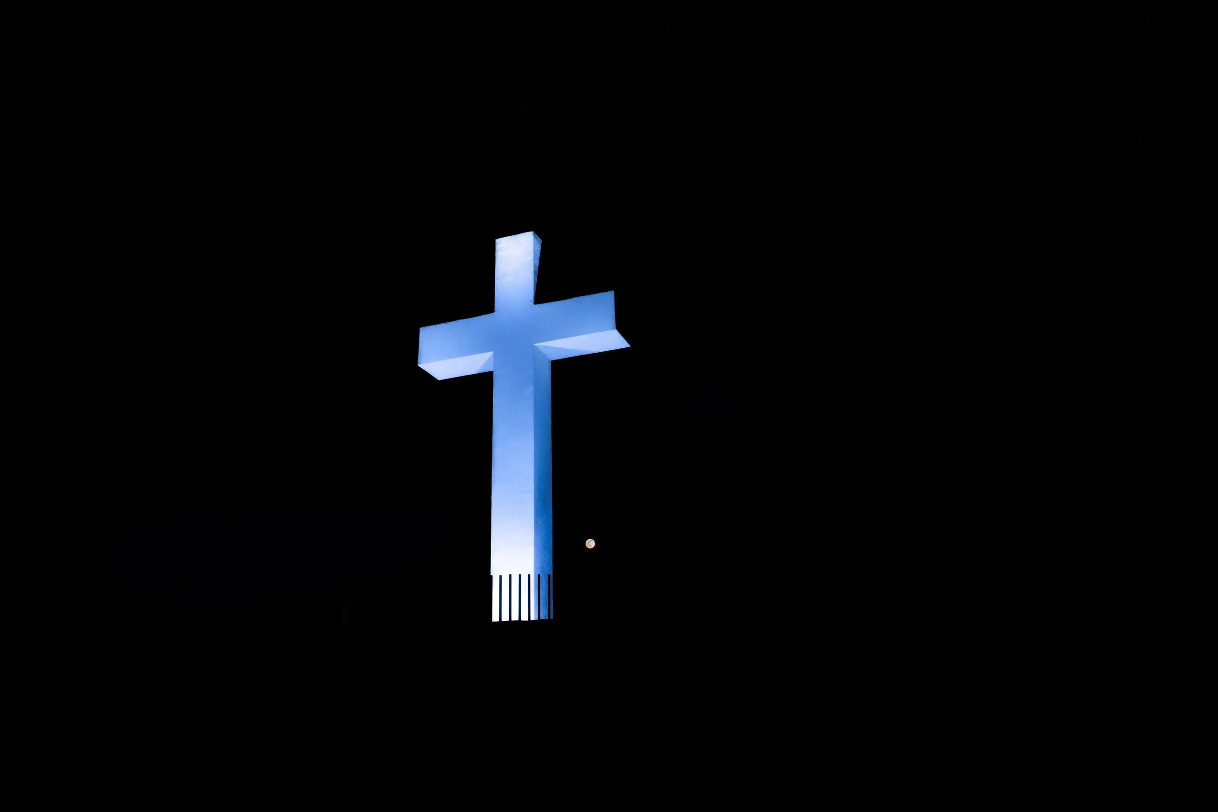 Illuminated blue cross at night with a small visible moon in the background.