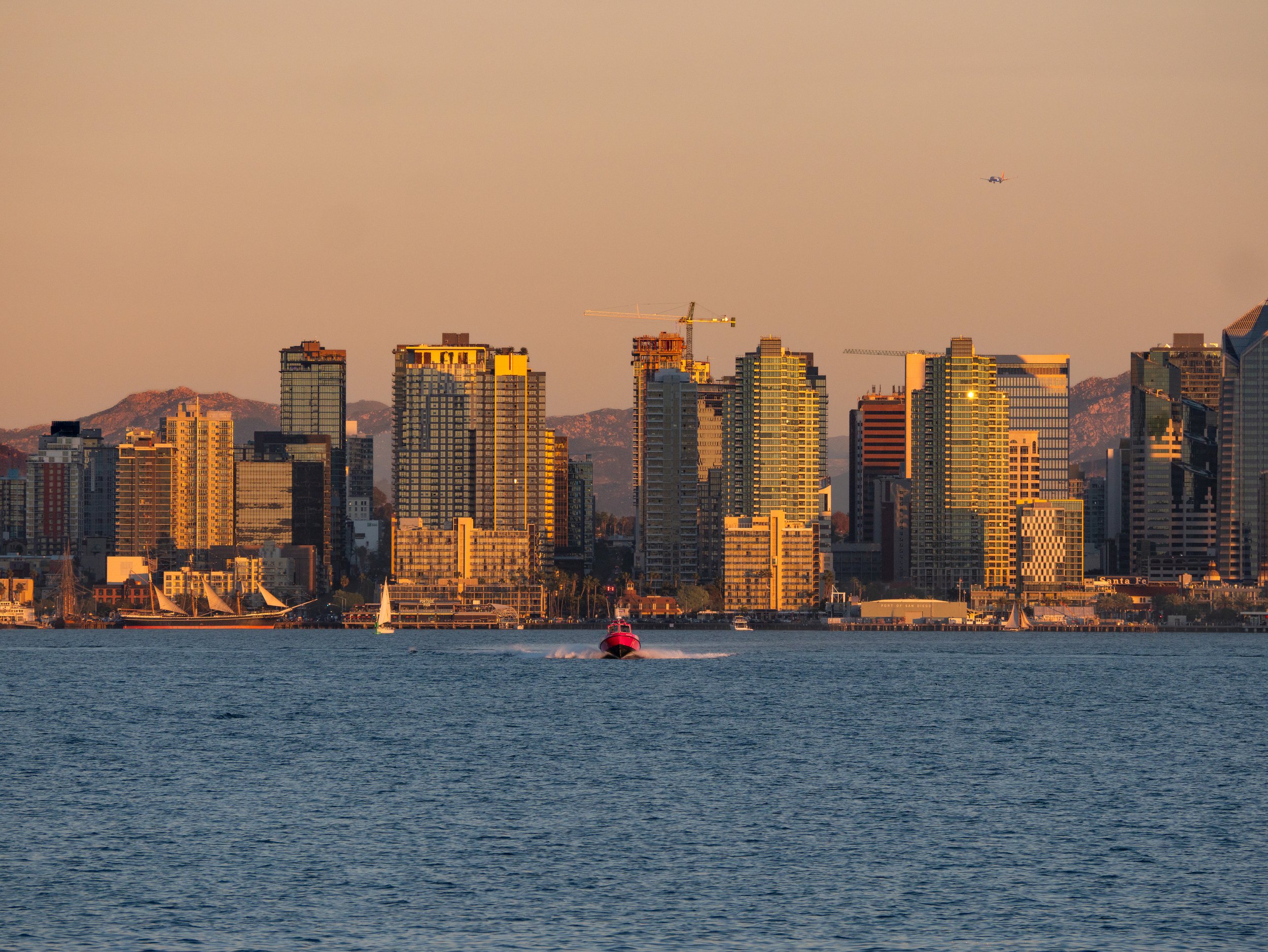 City skyline with tall buildings and mountains in the background, a boat in the water, and a small plane flying overhead during sunset.