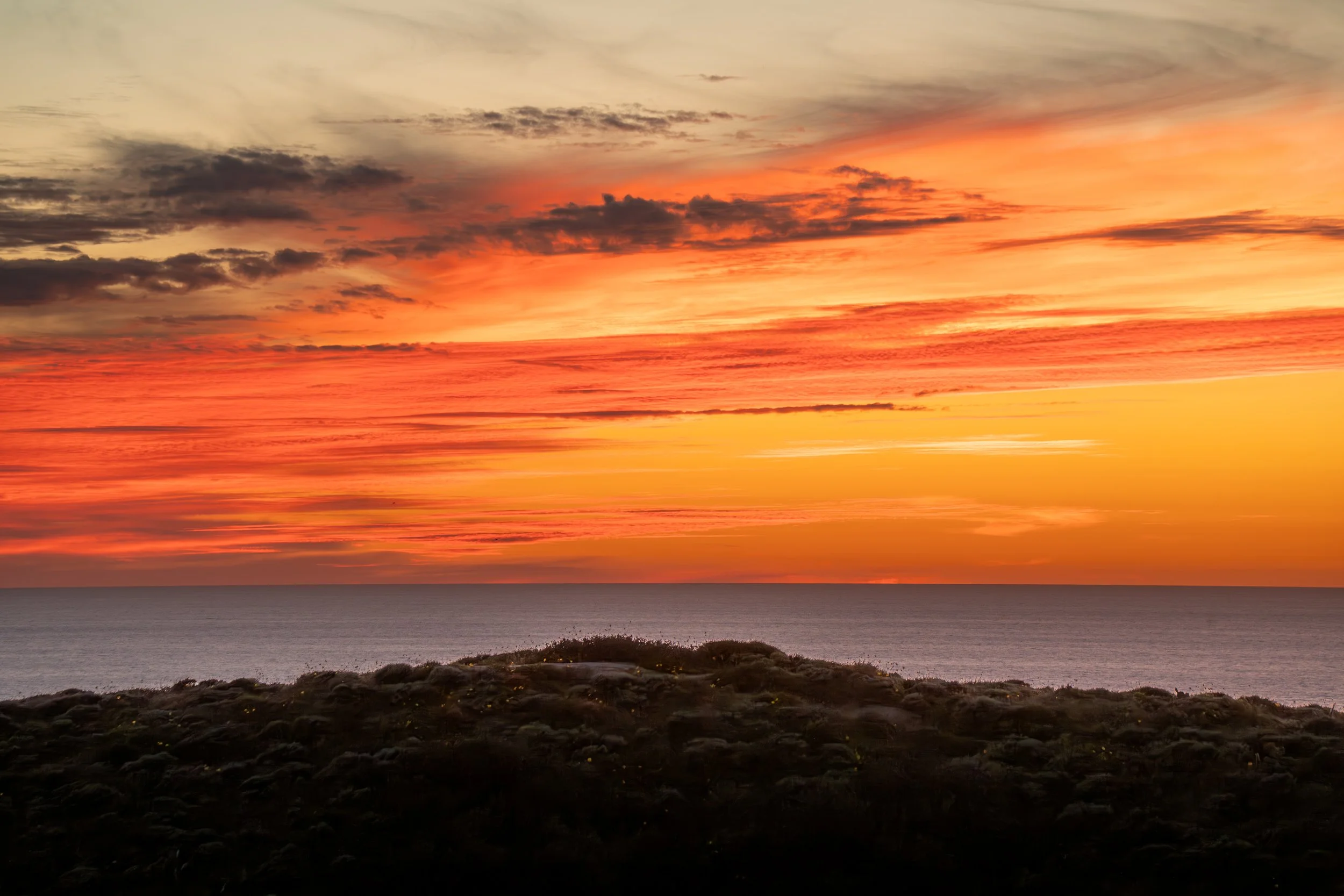 A sunset over the ocean with a dark rocky shoreline in the foreground, displaying vibrant orange, pink, and yellow colors across the sky and clouds.