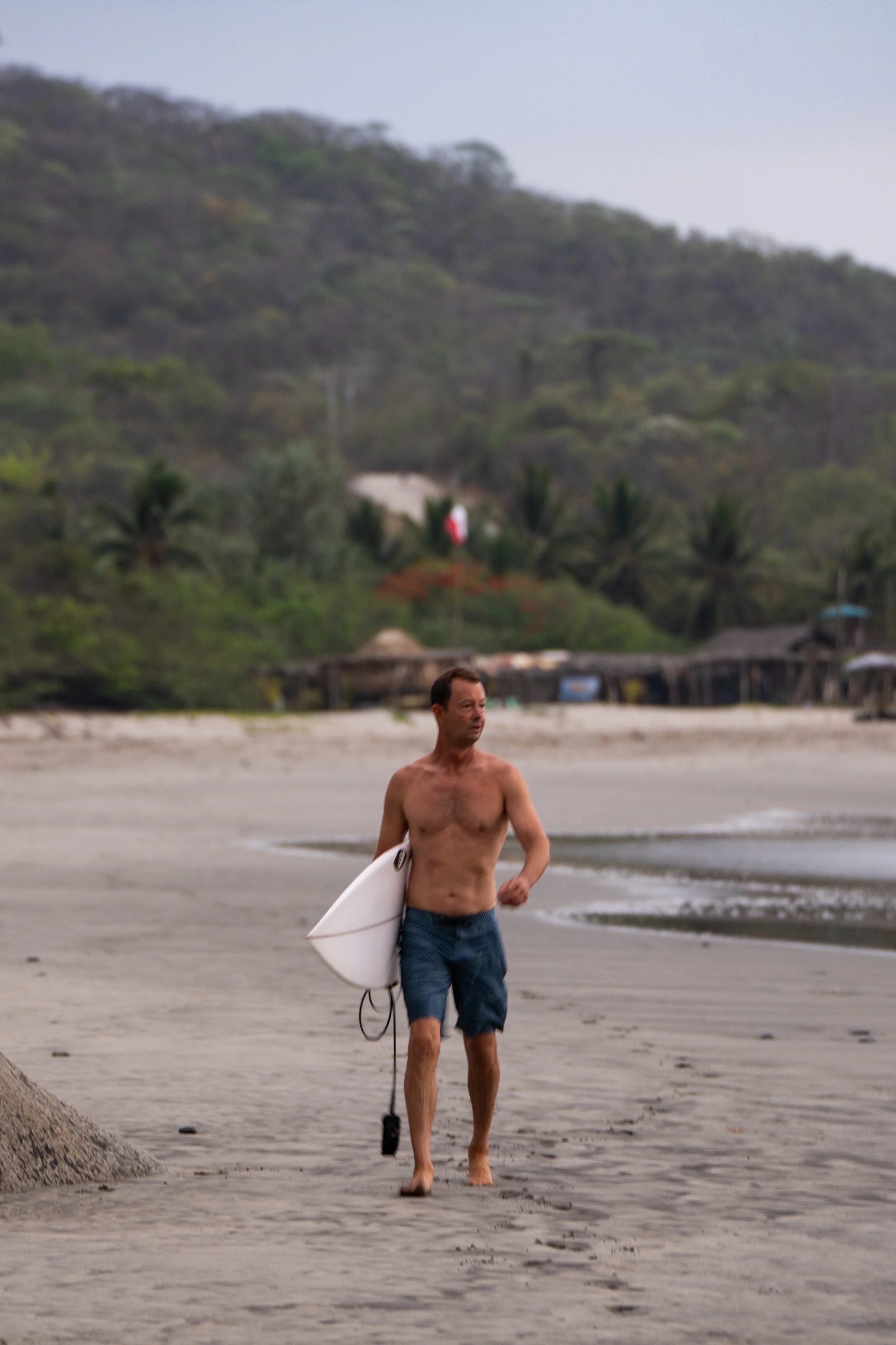 A shirtless man walking on a beach holding a surfboard in his right hand, with a scenic backdrop of lush green hills and trees.