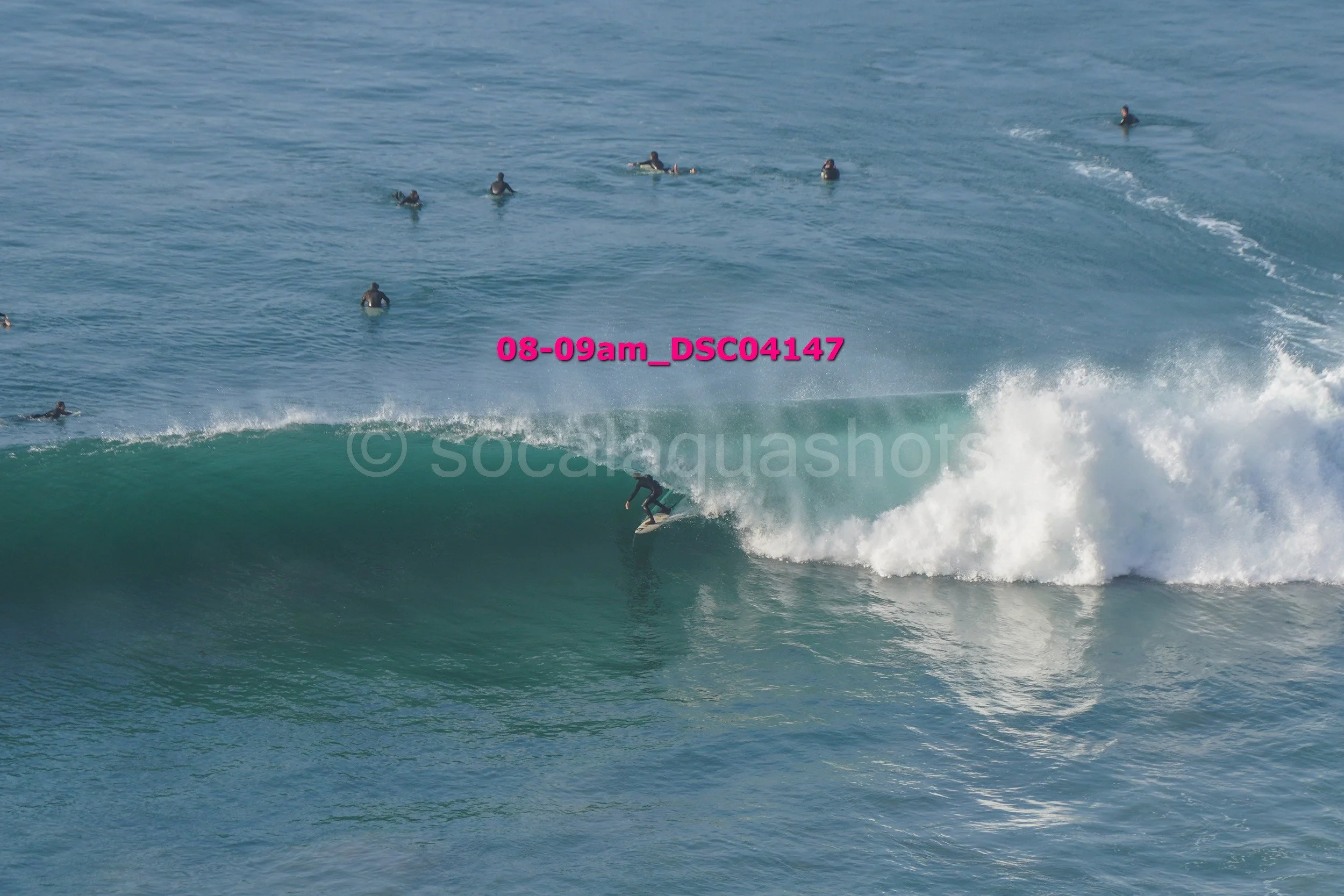 A surfer riding a large wave with several surfers watching from the water in the background.