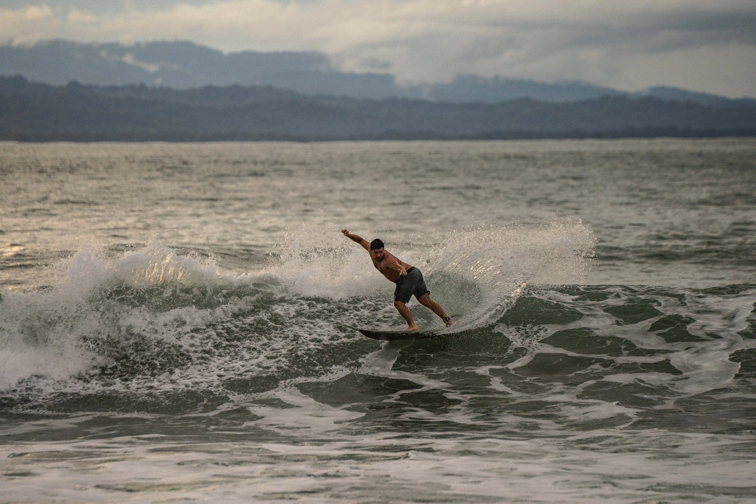A man surfing on a wave in the ocean with a mountain range and cloudy sky in the background.