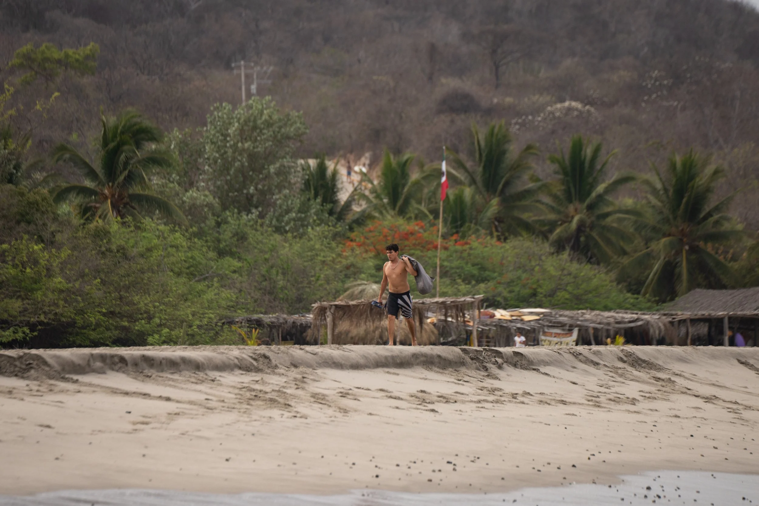 A young man walks along a beach carrying a jacket and a water bottle, with a village and trees in the background.