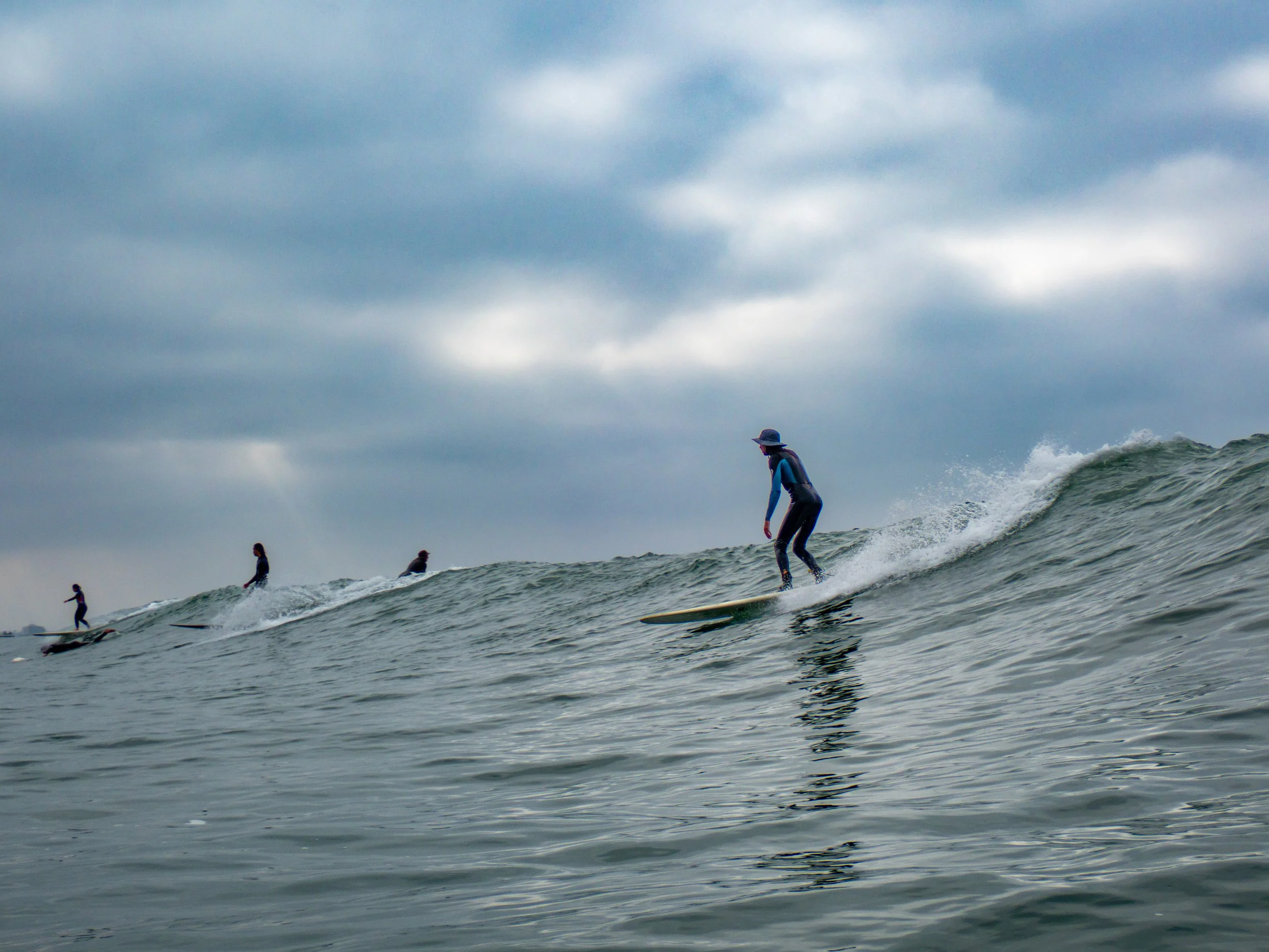 A person wearing a hat and wetsuit surfboarding on a wave with other surfers in the background under a cloudy sky.