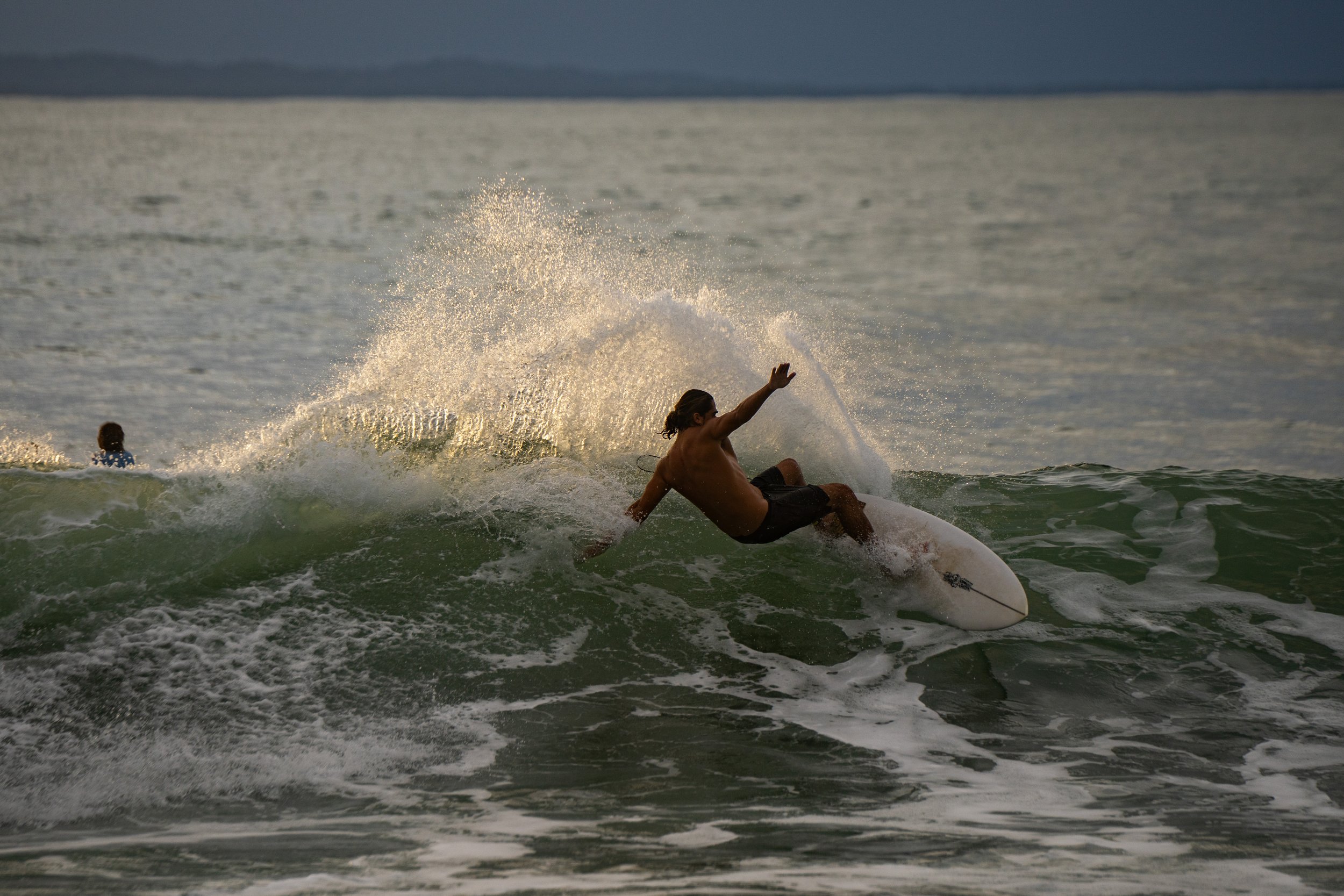 A person surfing on a wave in the ocean during sunset, with another person in the water in the background.
