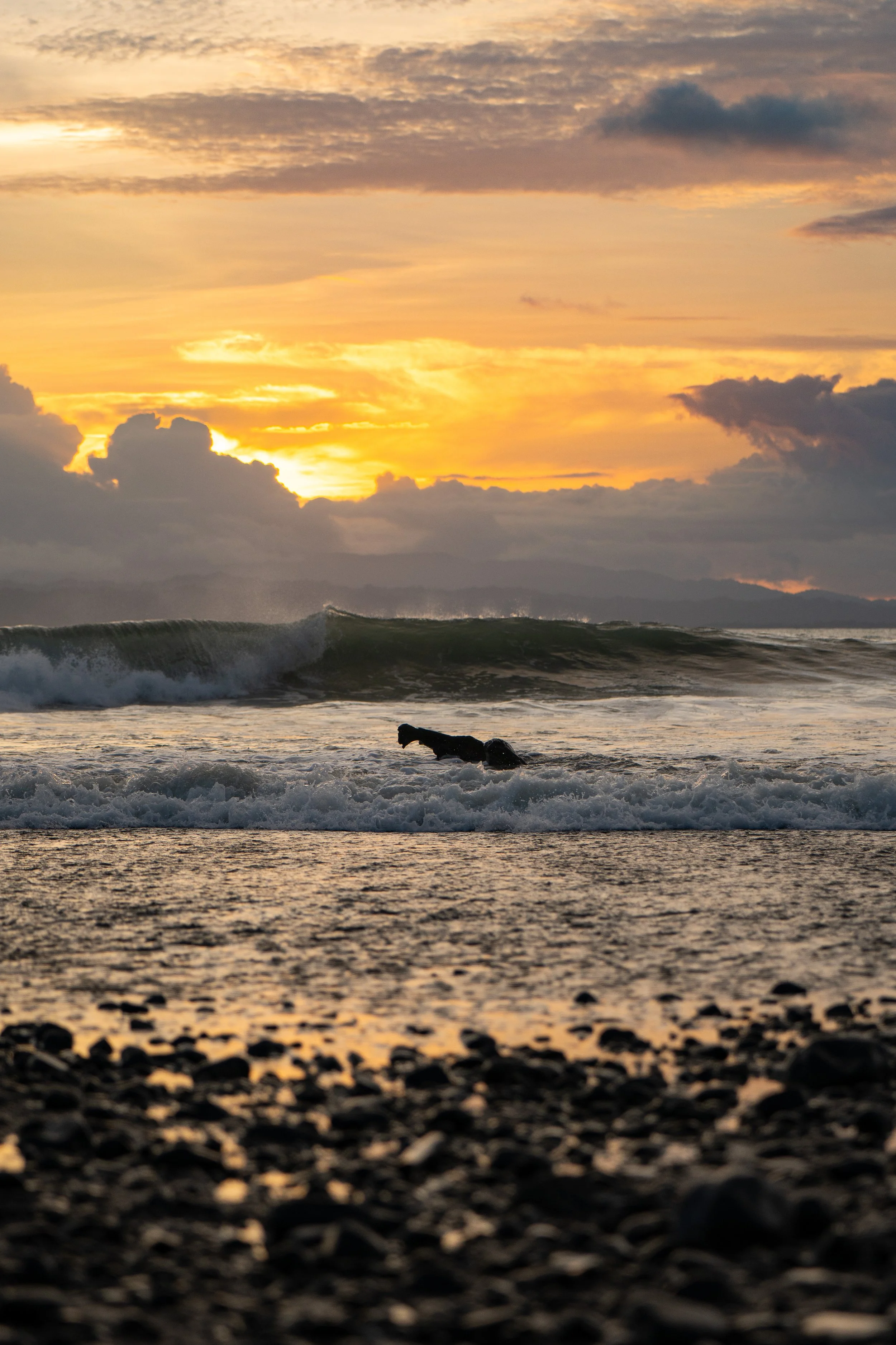 Sunset over the ocean with clouds, waves, and a rocky shoreline in the foreground.
