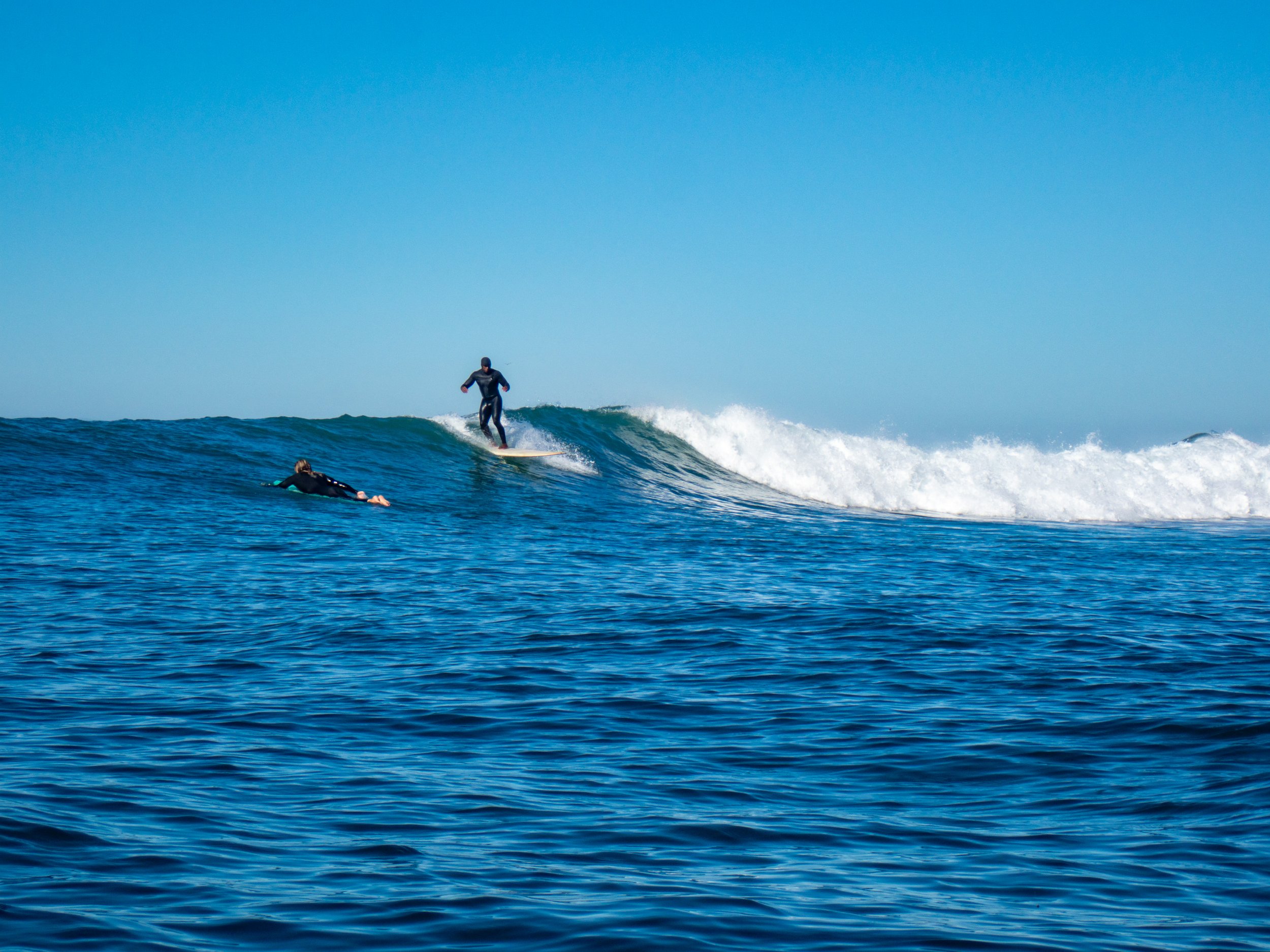 A person surfing on a wave with another person floating nearby in the ocean on a clear, sunny day.