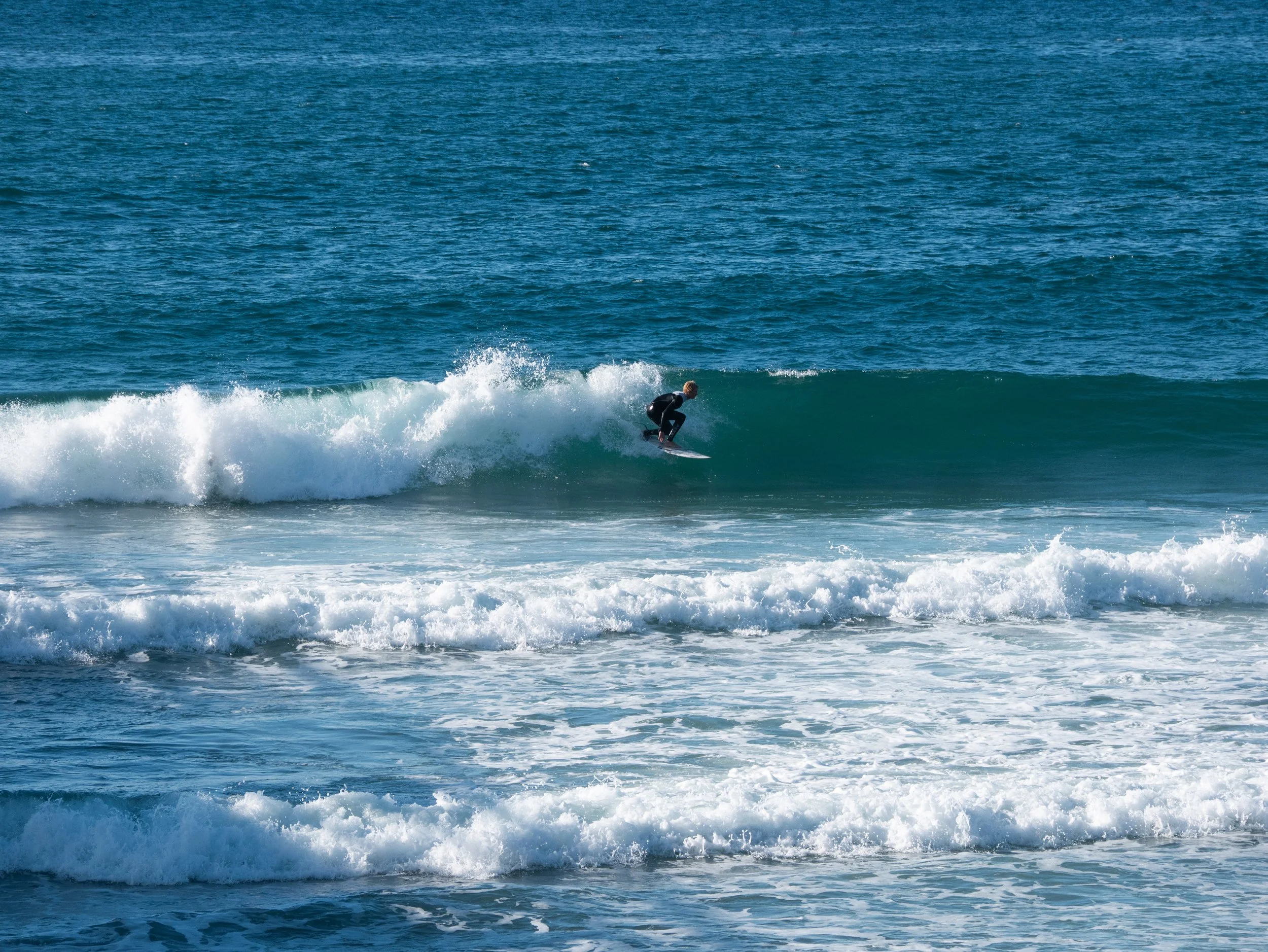 A person surfing on a small wave in the ocean, wearing a black wetsuit.