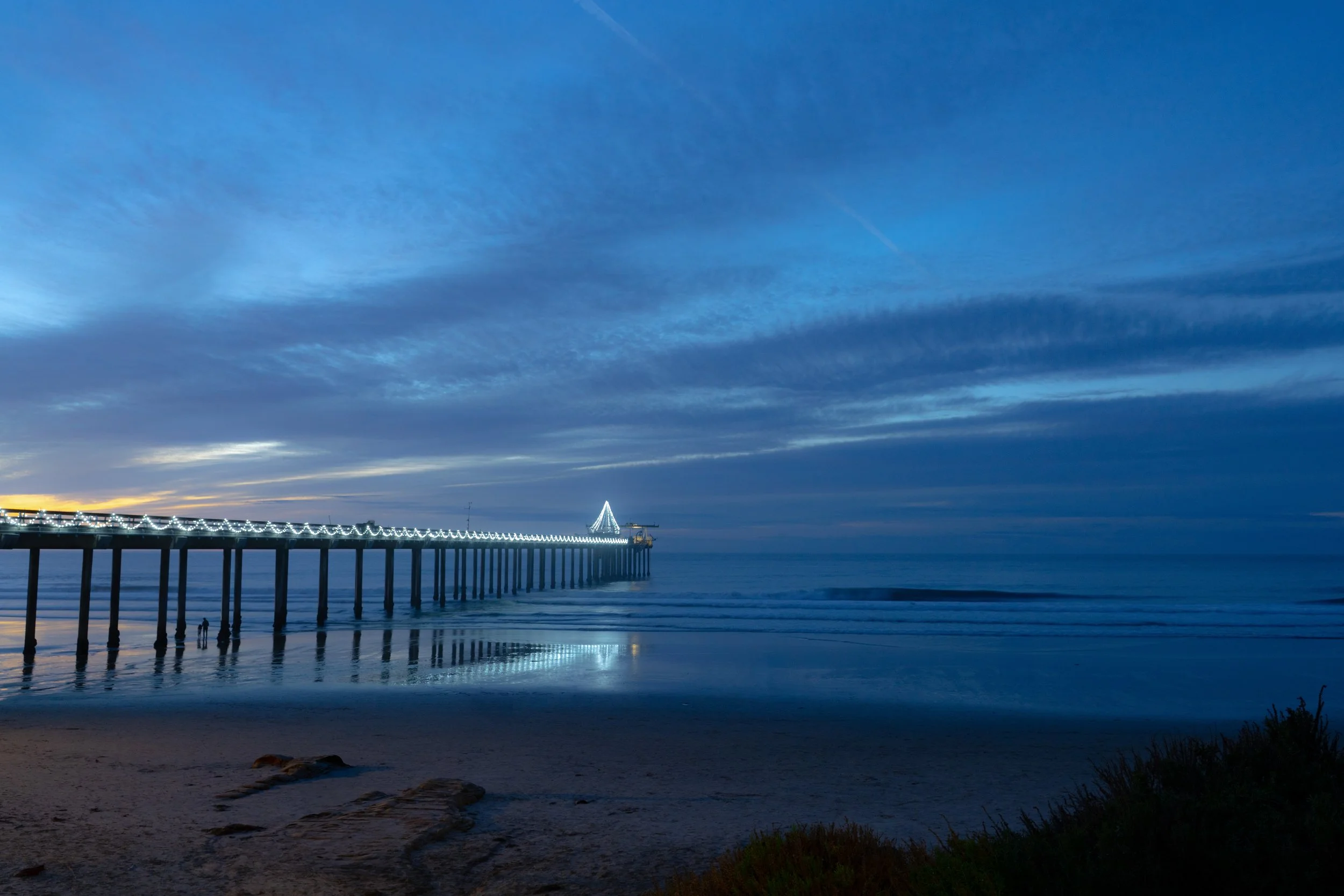 A long pier with colorful lights extending into the ocean at dusk, with a cloudy sky overhead and a sandy beach in the foreground.