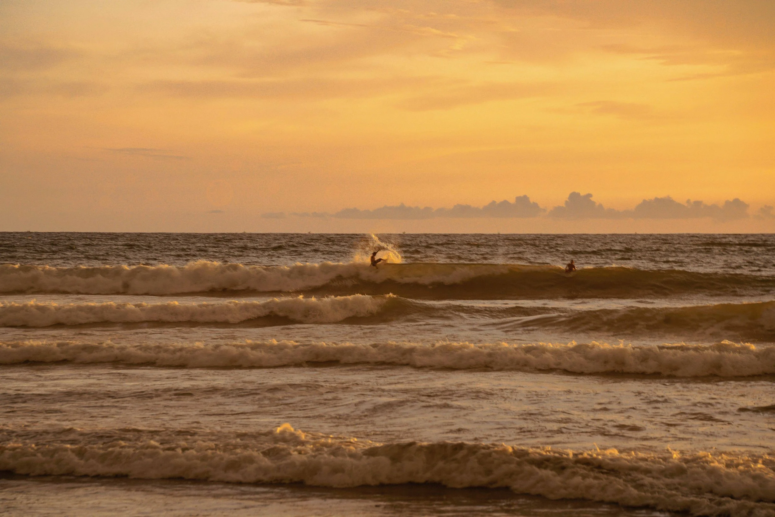 Sunset over the ocean with waves and surfers in the water.