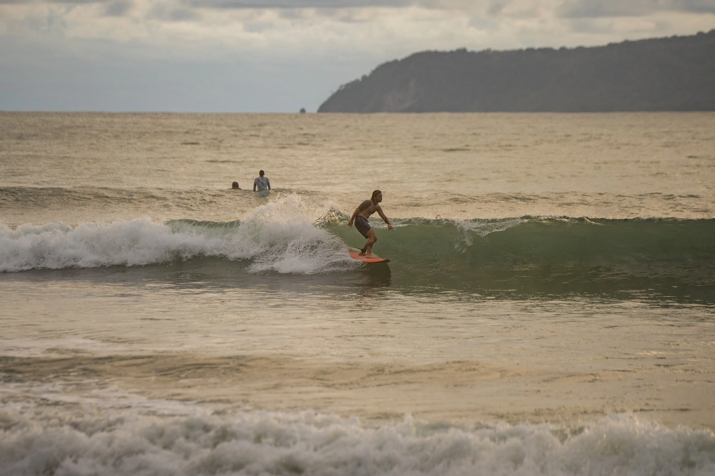 A man surfing on a small wave in the ocean with two other people swimming behind him. A large landmass and cloudy sky are visible in the background.