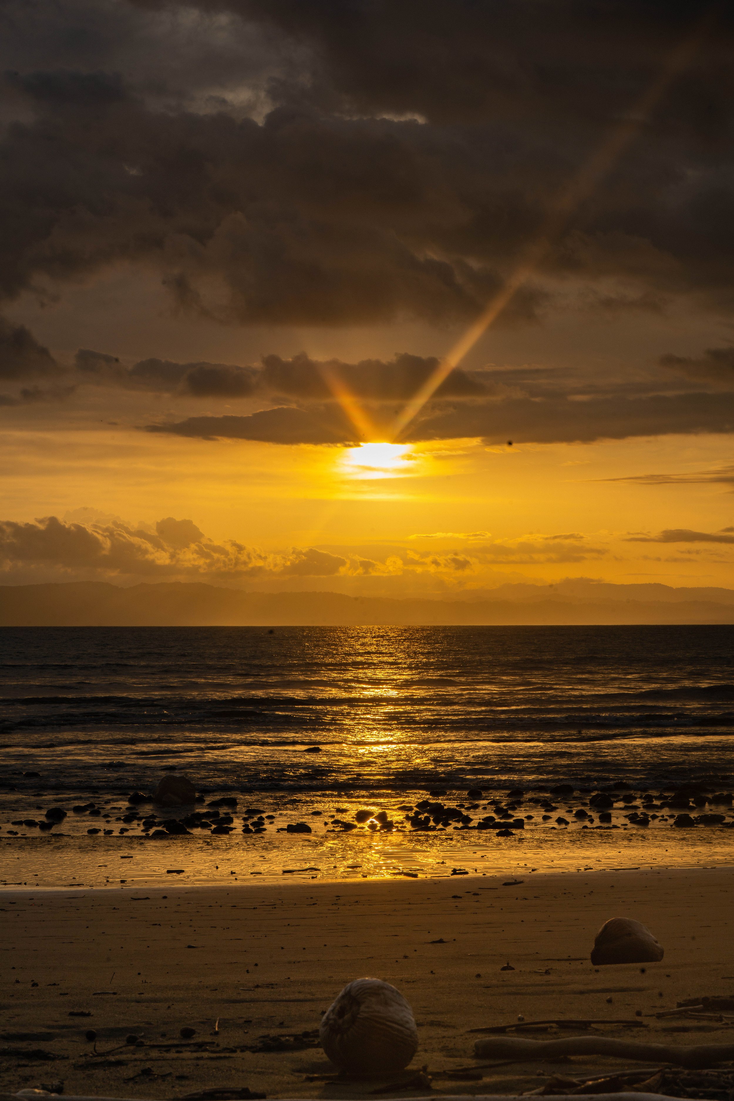 Sunset over the ocean with a cloudy sky and rocks on the sandy beach in the foreground.