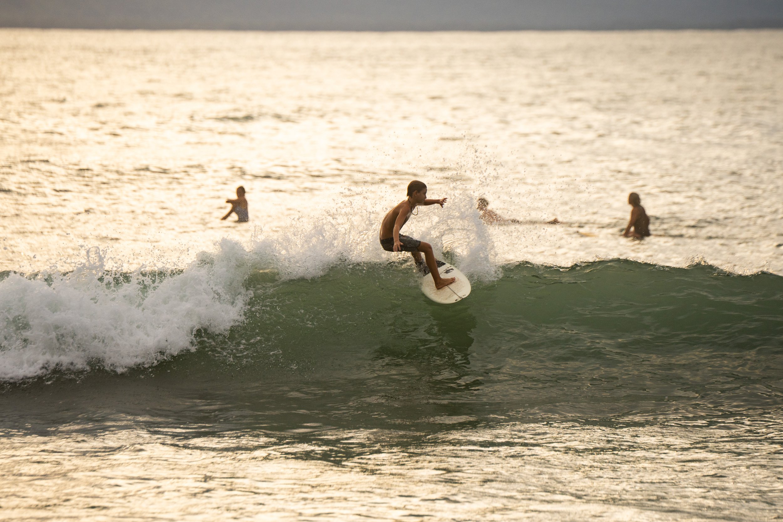 A person surfing on a wave in the ocean during sunset, with other people swimming in the water nearby.