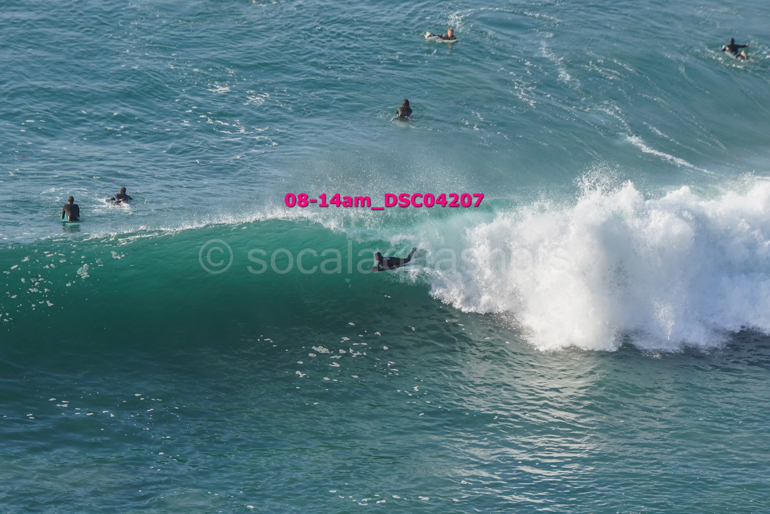 A person surfing on a wave while others are swimming or floating in the water nearby.