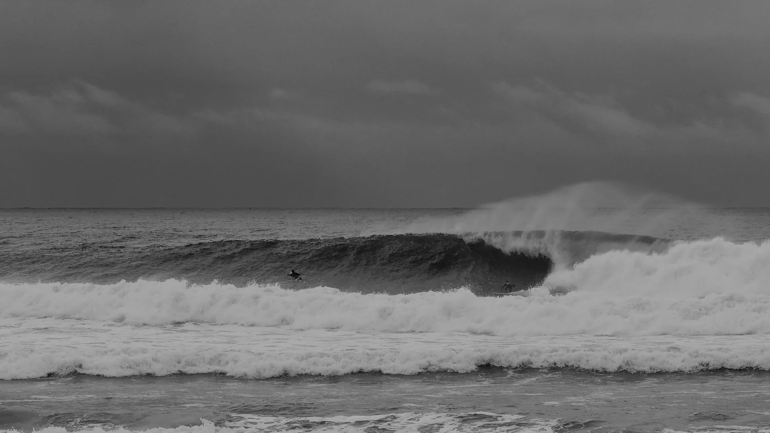 Black-and-white surf photograph of surfers riding a large ocean wave.