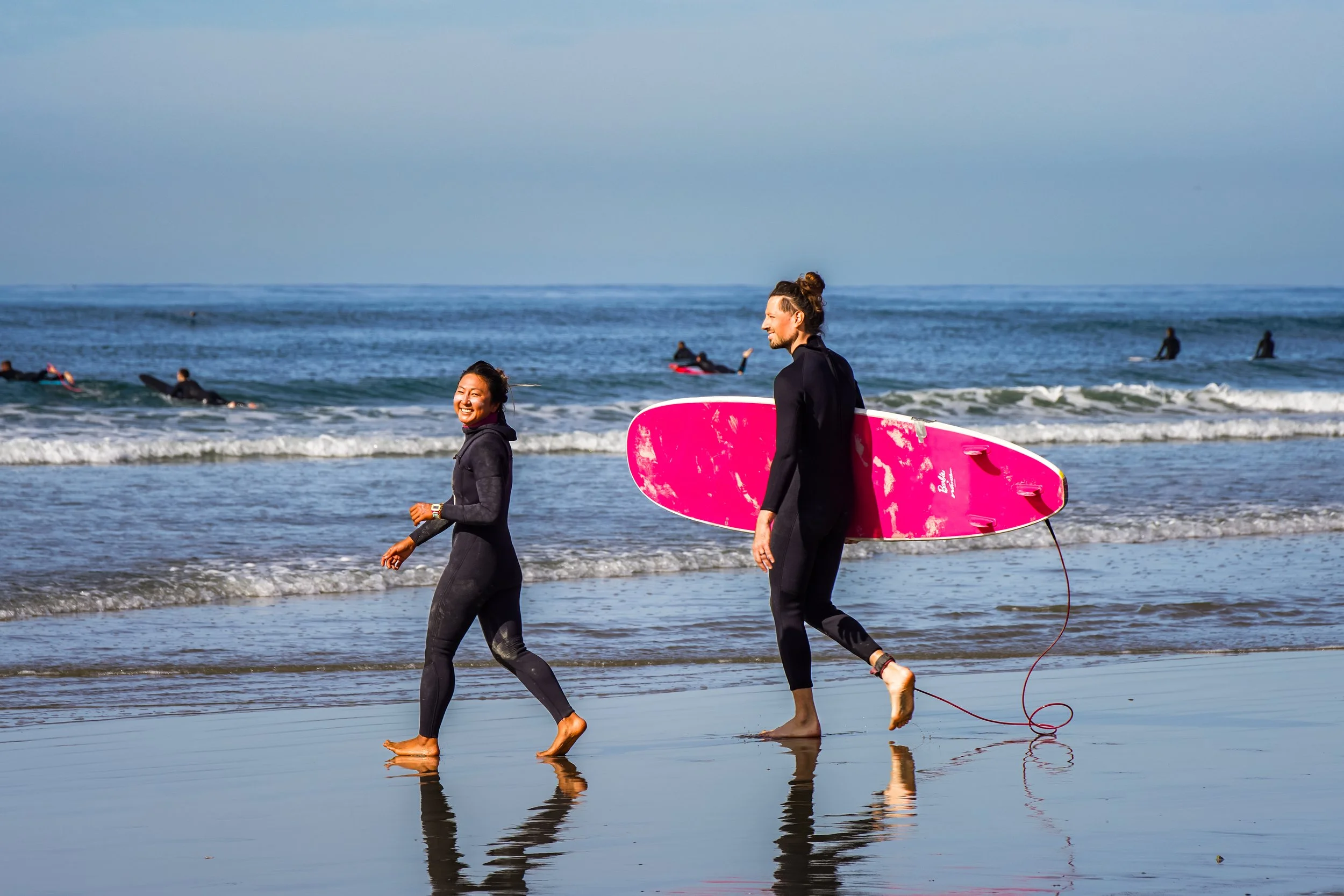 Two people in wetsuits walking on the beach with a pink surfboard. The person on the right carries the surfboard, and the one on the left looks back smiling. The ocean with several surfers in the background and a cloudy sky is visible.