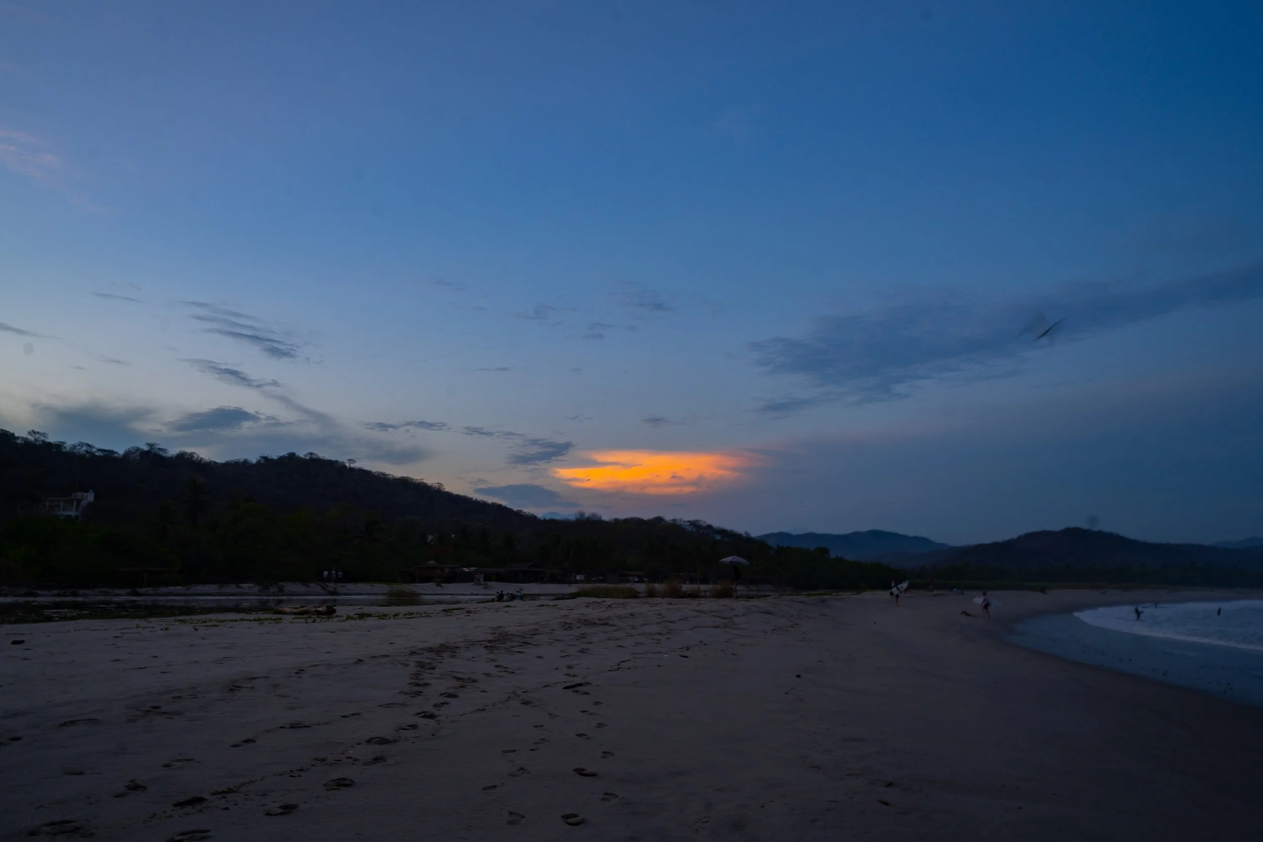 Sunset over a beach with a few people walking along the shoreline, trees and hills in the background, and a clear sky with some clouds.
