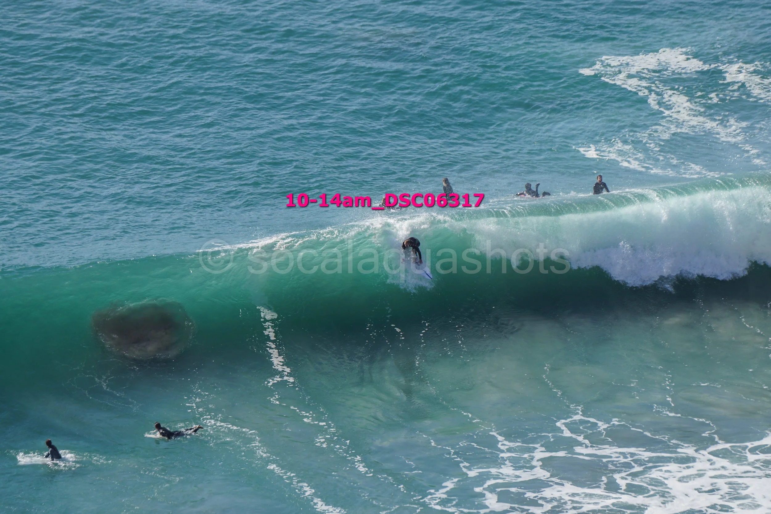 Surfer riding a wave with several people in the water and a large rock visible beneath the wave.