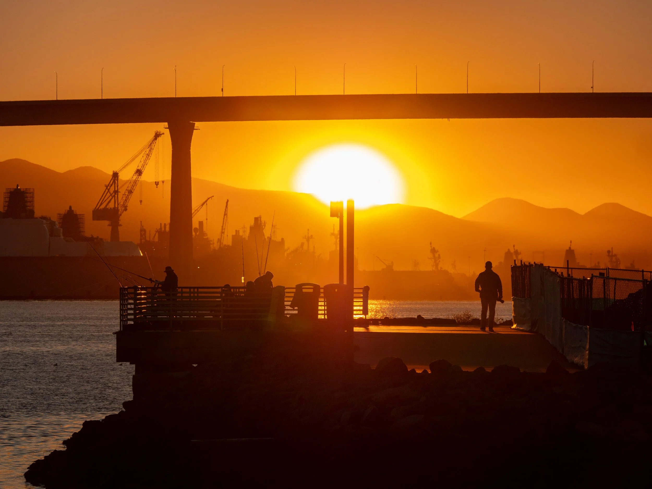 Silhouetted people fishing on a pier at sunset with an industrial harbor, cranes, and mountains in the background.