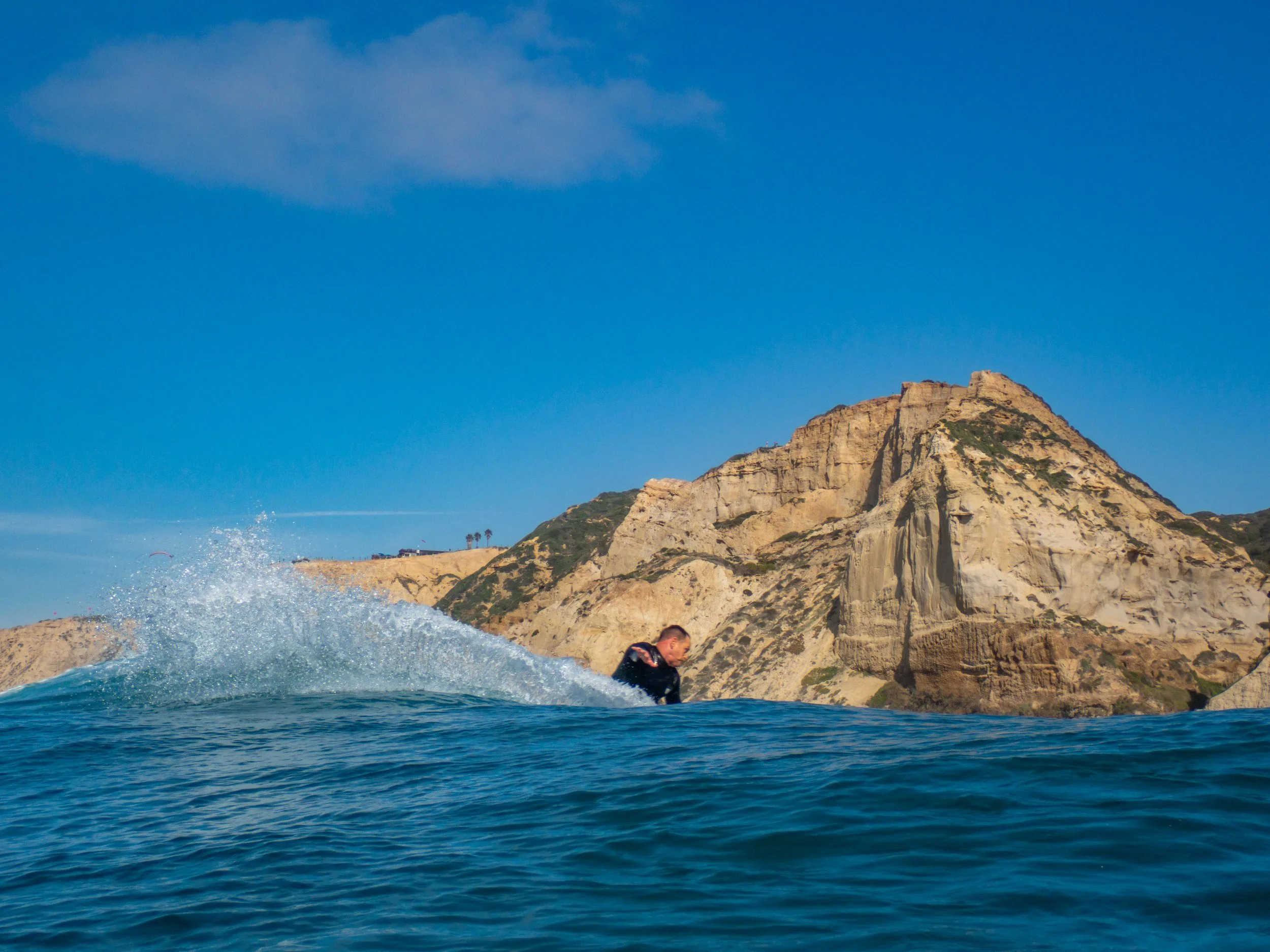 A person surfing in the ocean near rocky cliffs under a clear blue sky.