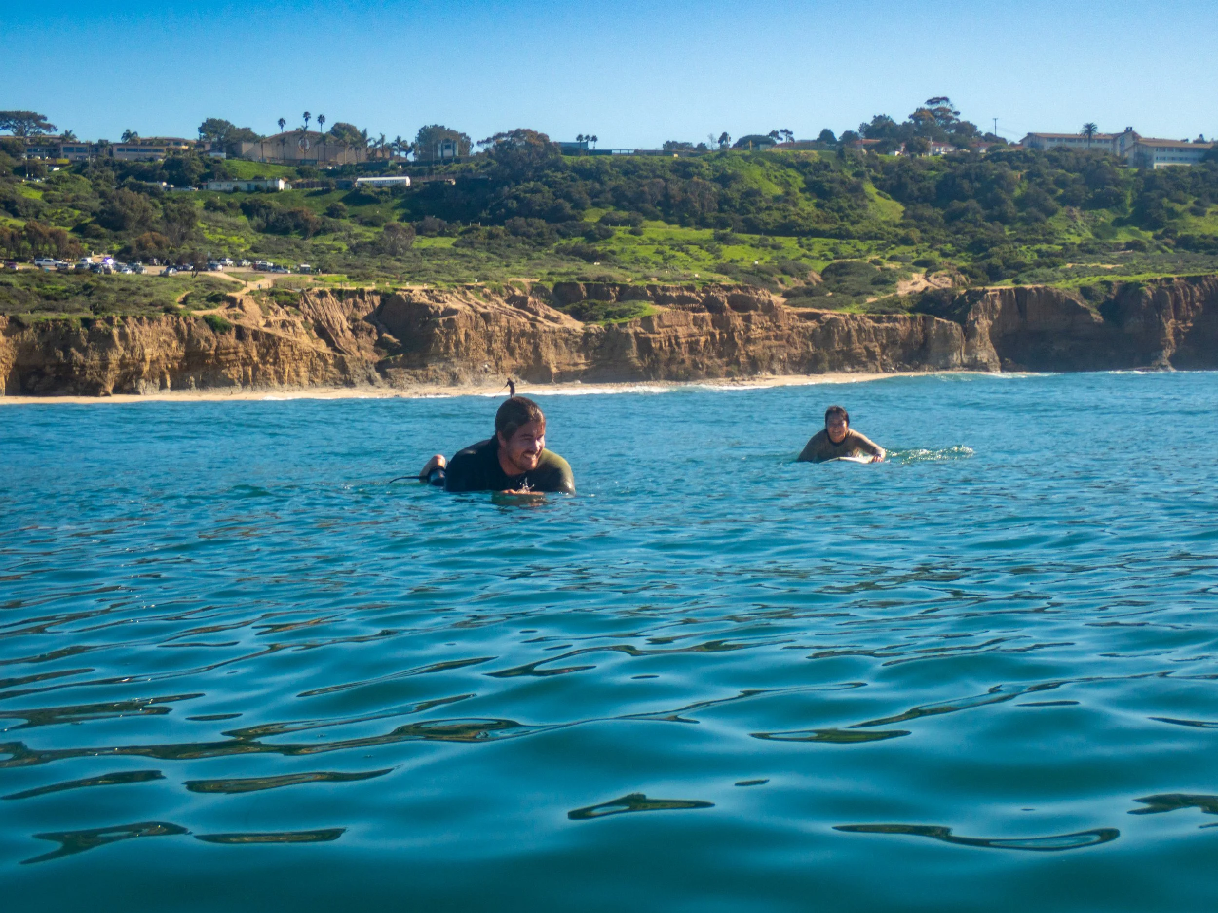 Two people in the ocean on surfboards with a rocky shoreline and green hills in the background.
