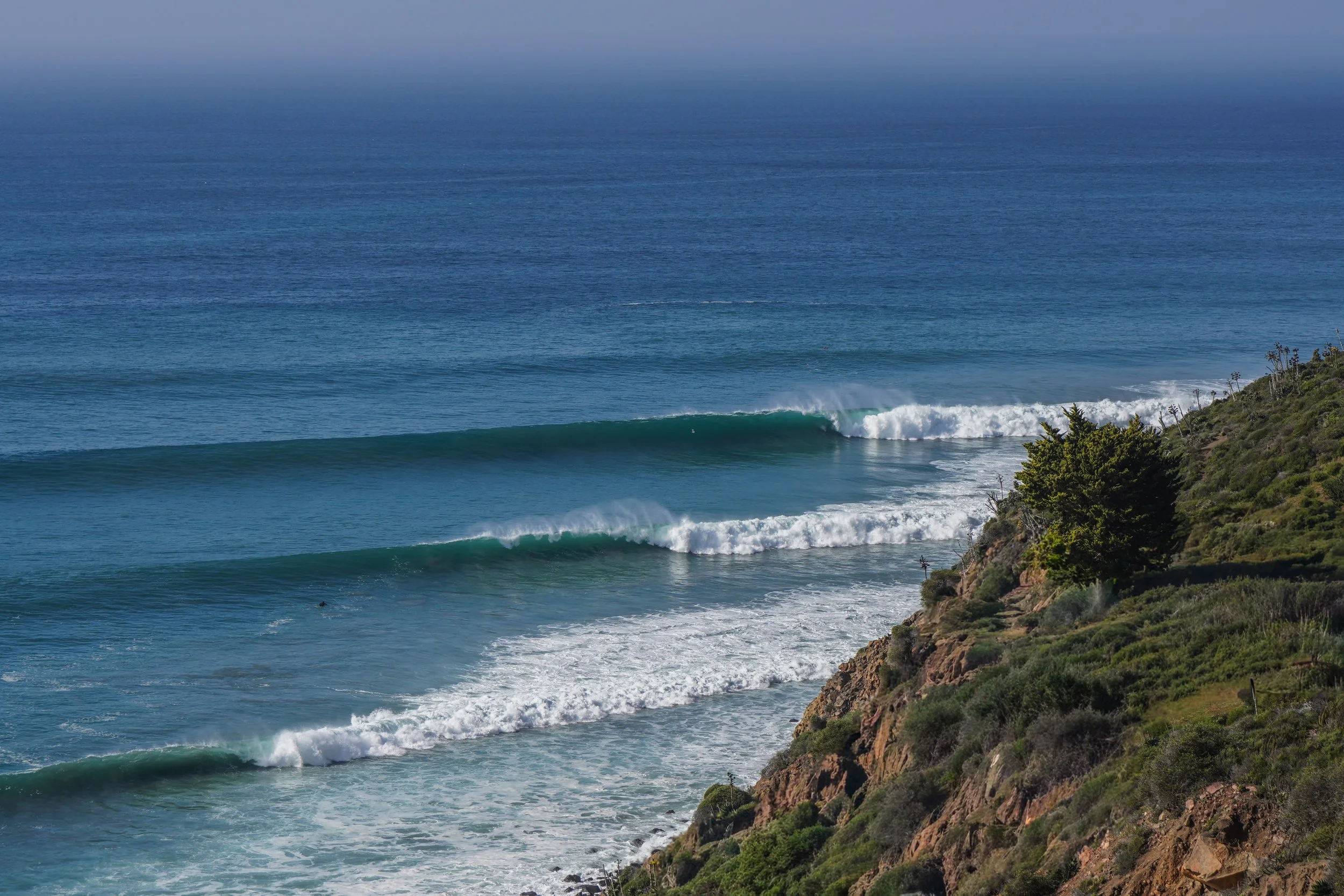 Ocean waves crashing along a rocky coastal shoreline with green bushes and trees.