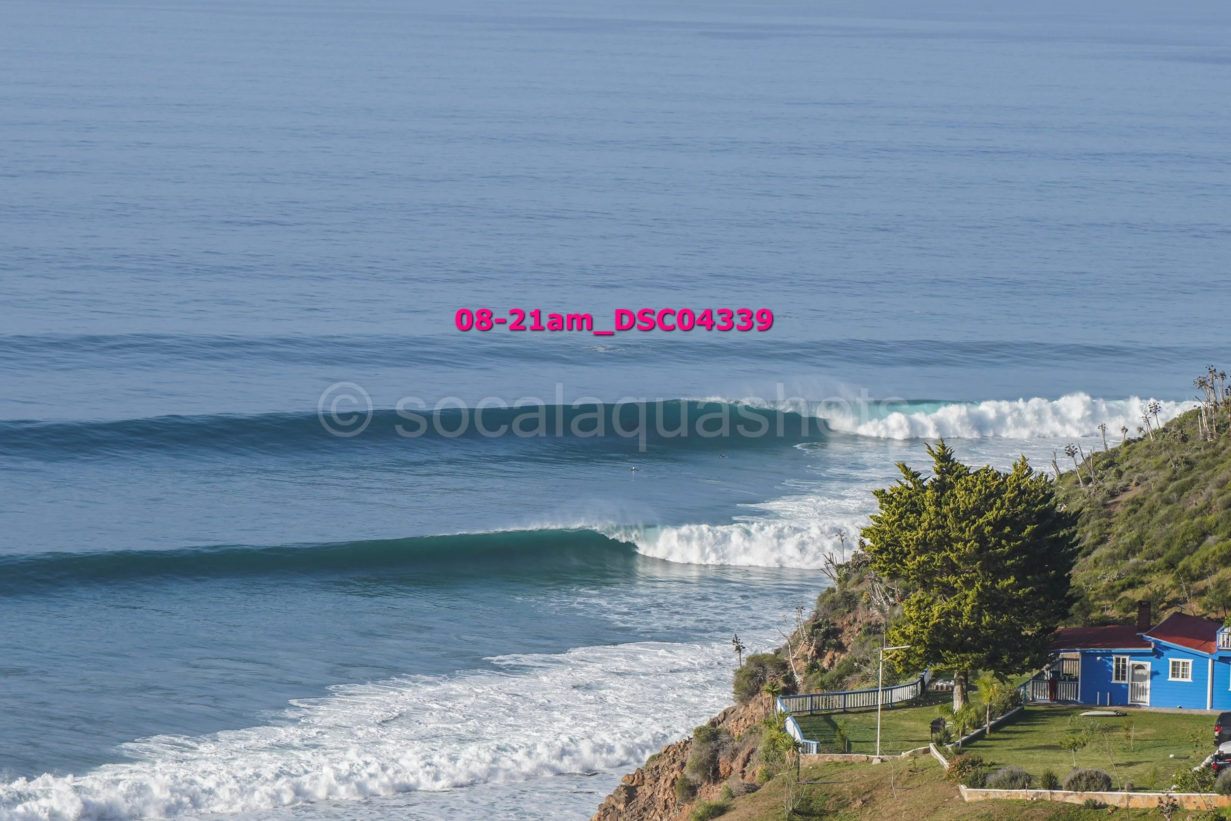 View of the ocean with waves breaking near a coastal beach, houses, and trees on the shore.