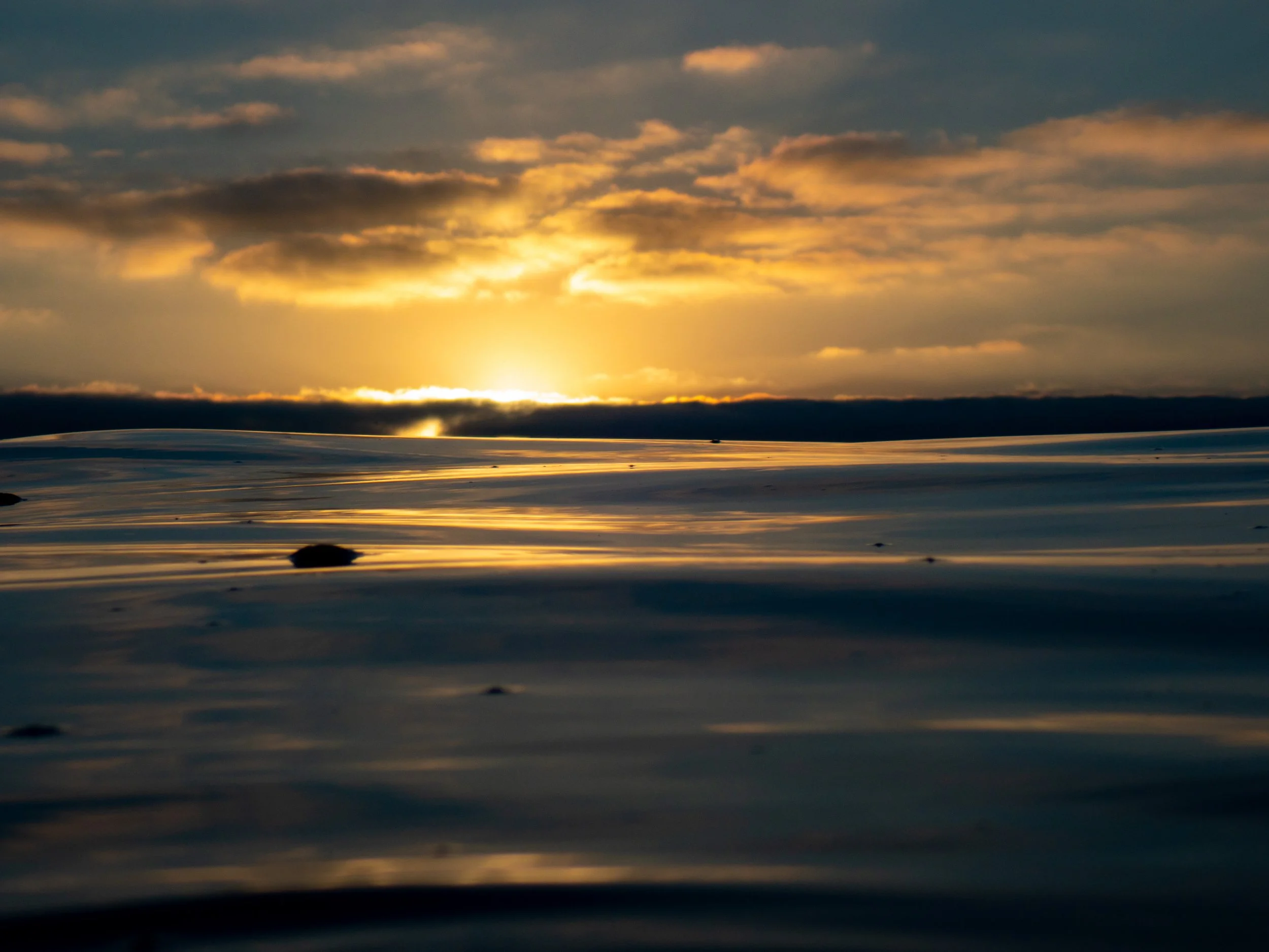 Sunset over the ocean with a cloudy sky and rippling water, some rocks in the foreground.