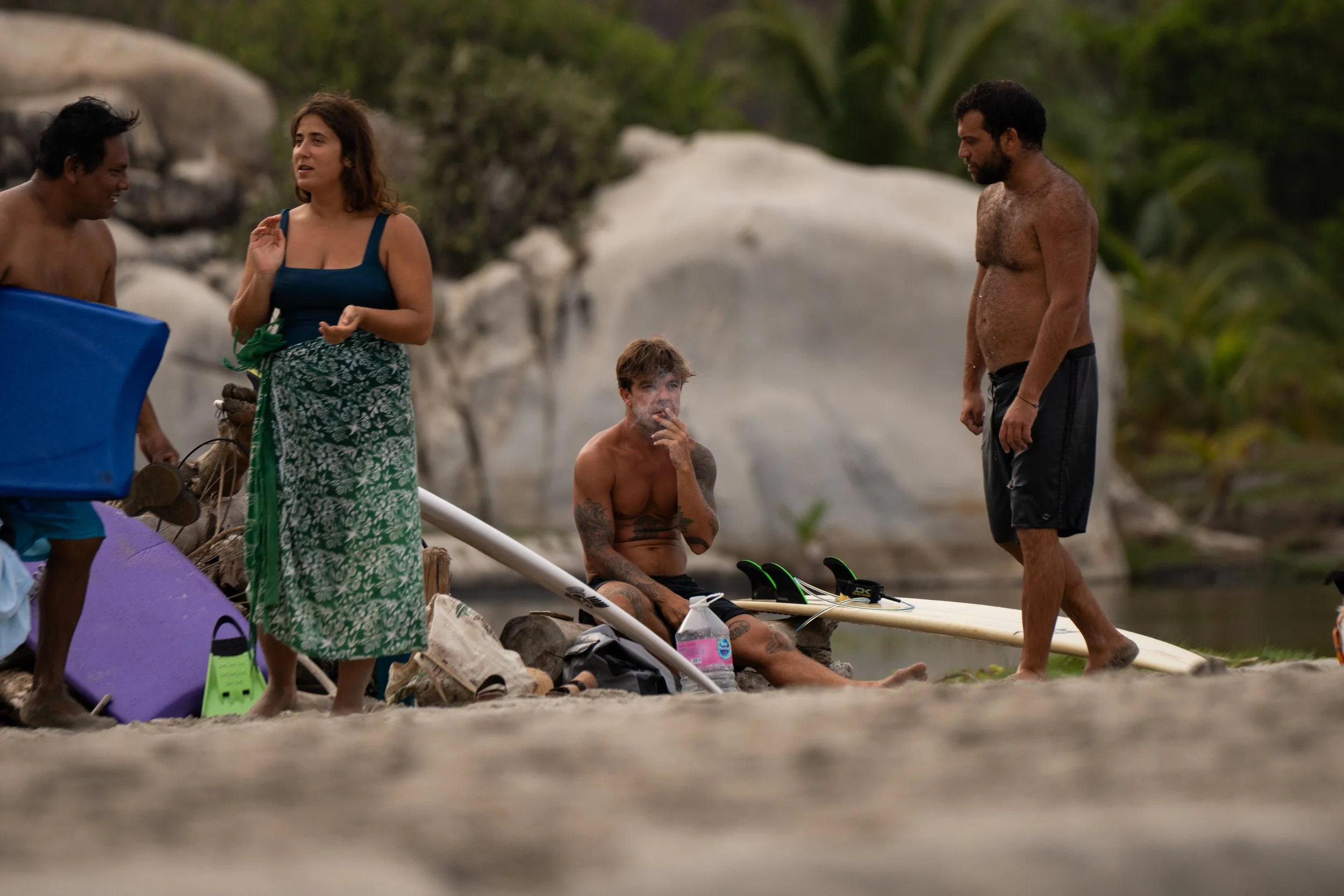 Four people on a beach, with one sitting and smoking, two standing shirtless, and one woman in a blue top and patterned skirt, with surfboards and gear around.