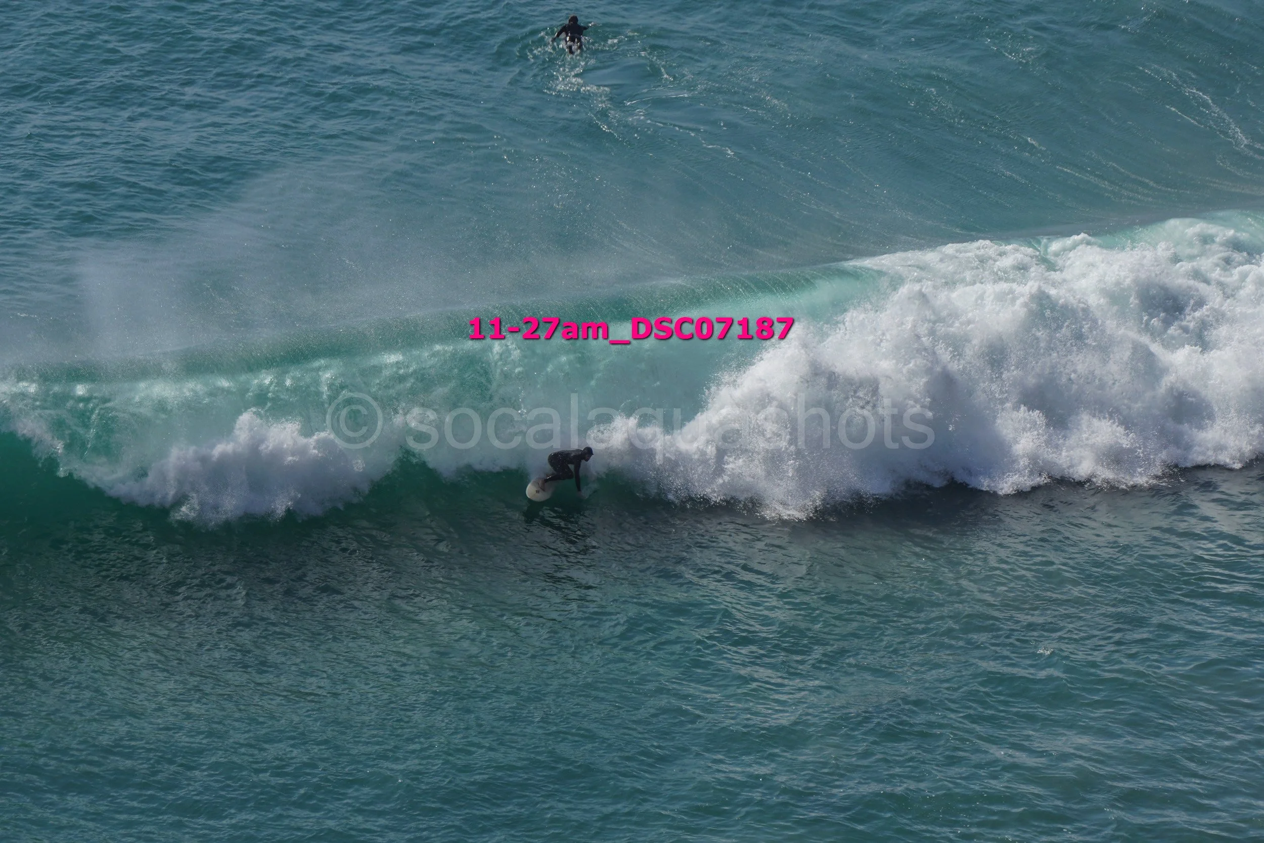 A surfer riding inside a large ocean wave with another surfer visible in the background.