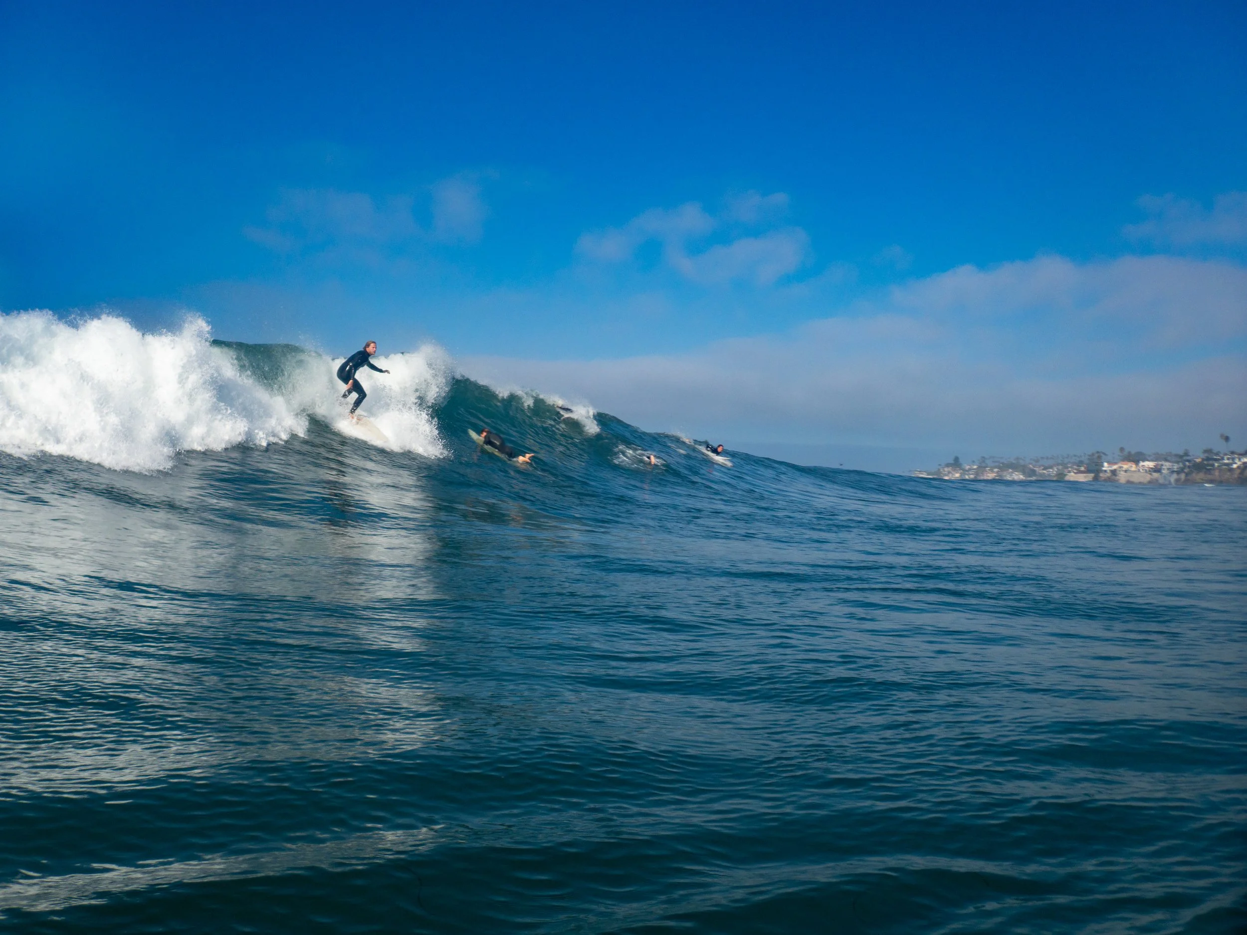 A person surfing on a wave in the ocean with other surfers in the water and a distant coastline under a blue sky.