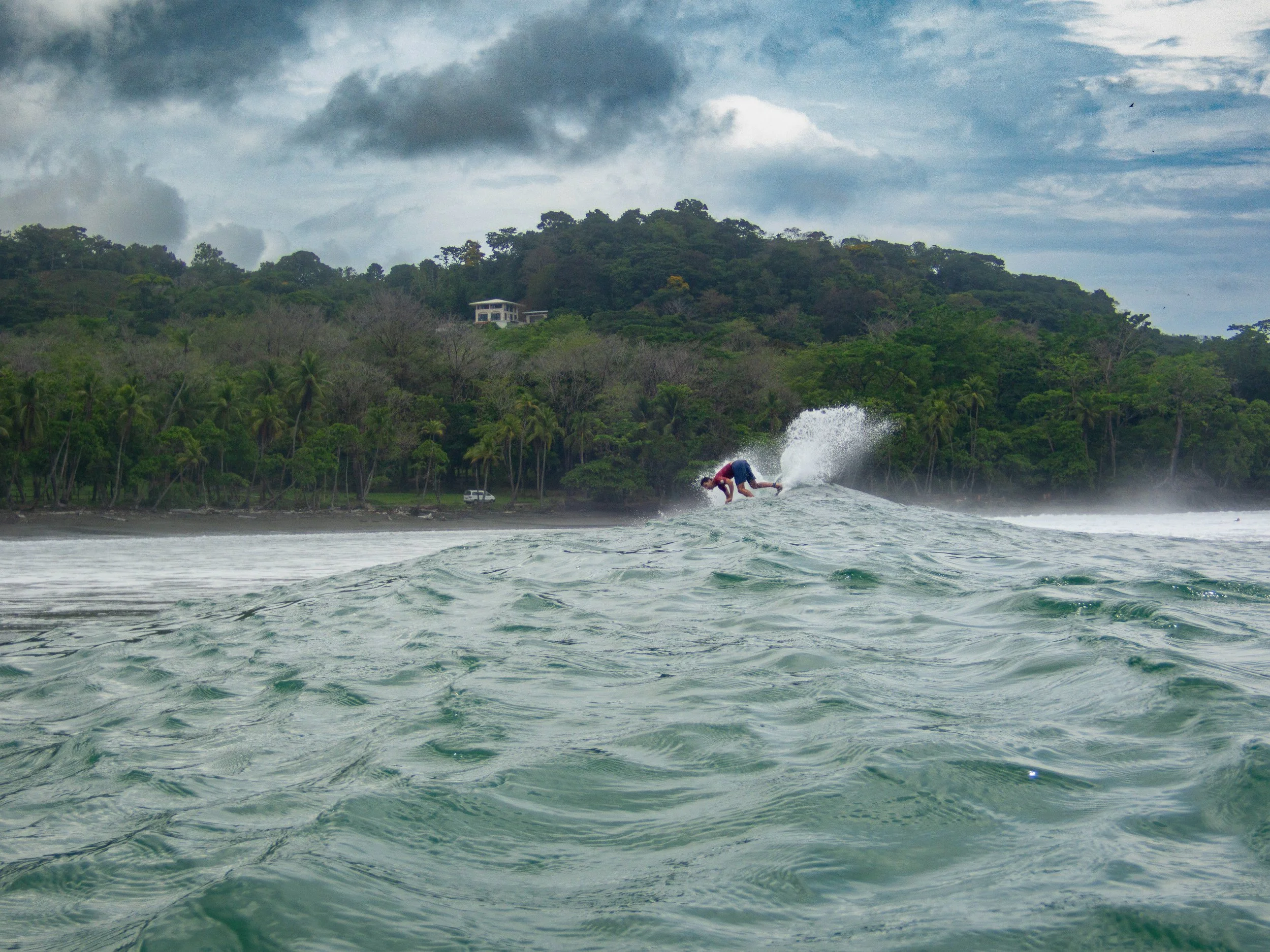 Person surfing on a wave with a tropical forest in the background under a cloudy sky.