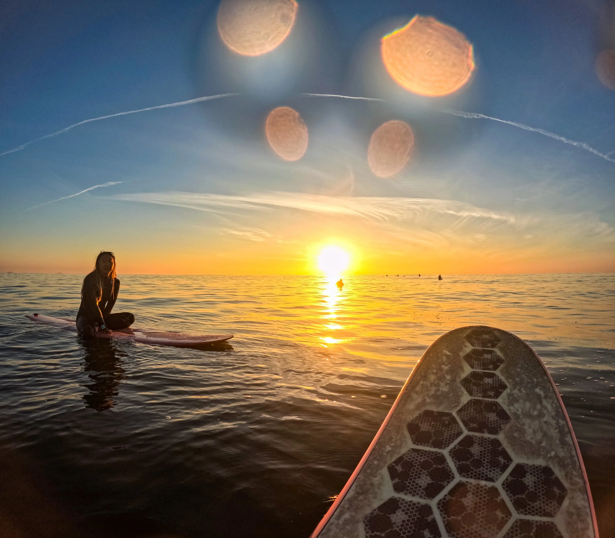 A person sitting on a paddleboard in the ocean during sunset, with the underside of another paddleboard in the foreground and a woman on a paddleboard in the distance. The sky is partly cloudy with the sun near the horizon.