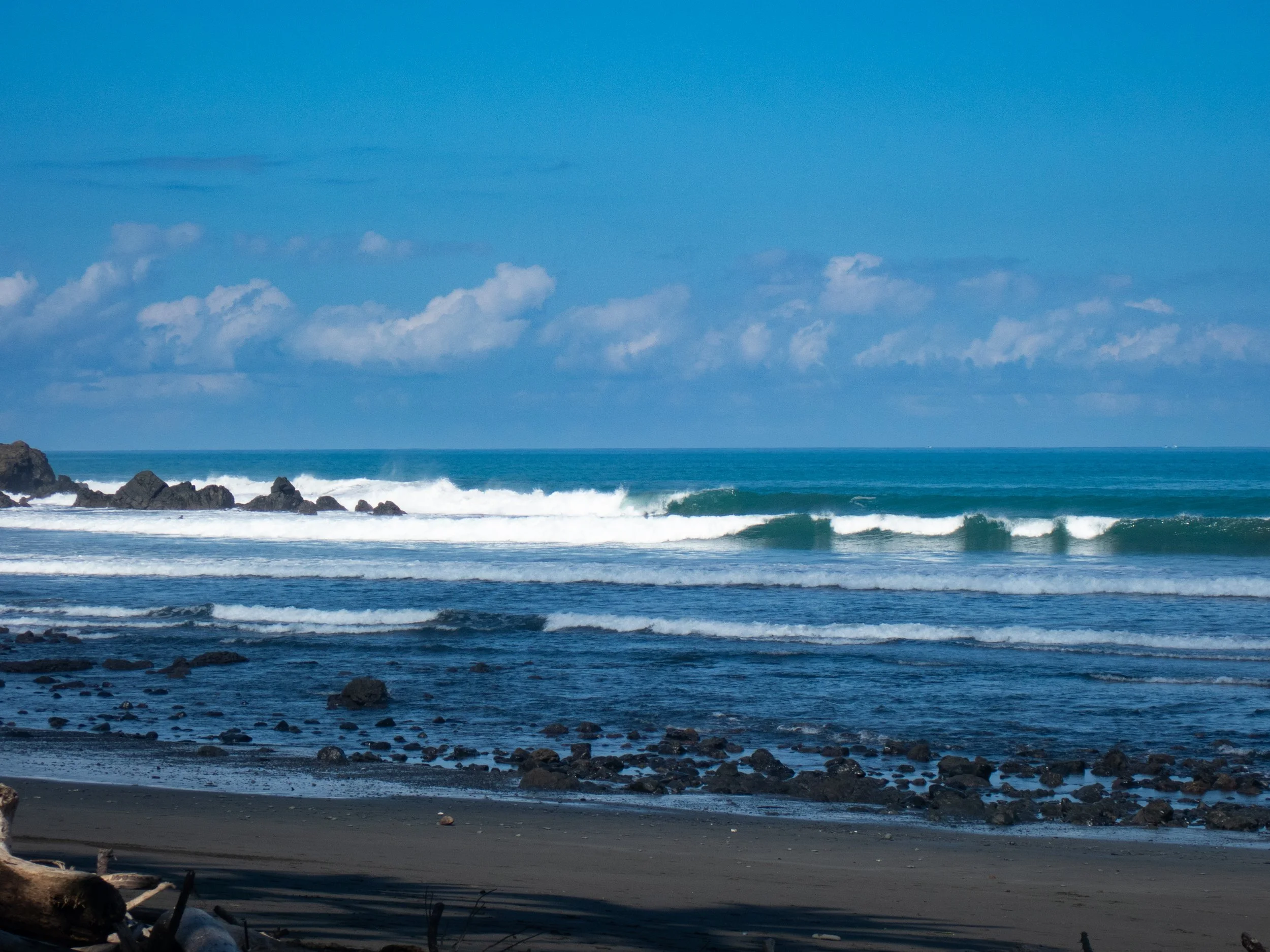 A scenic view of the ocean with waves crashing onto a rocky shoreline and a partly cloudy blue sky above.