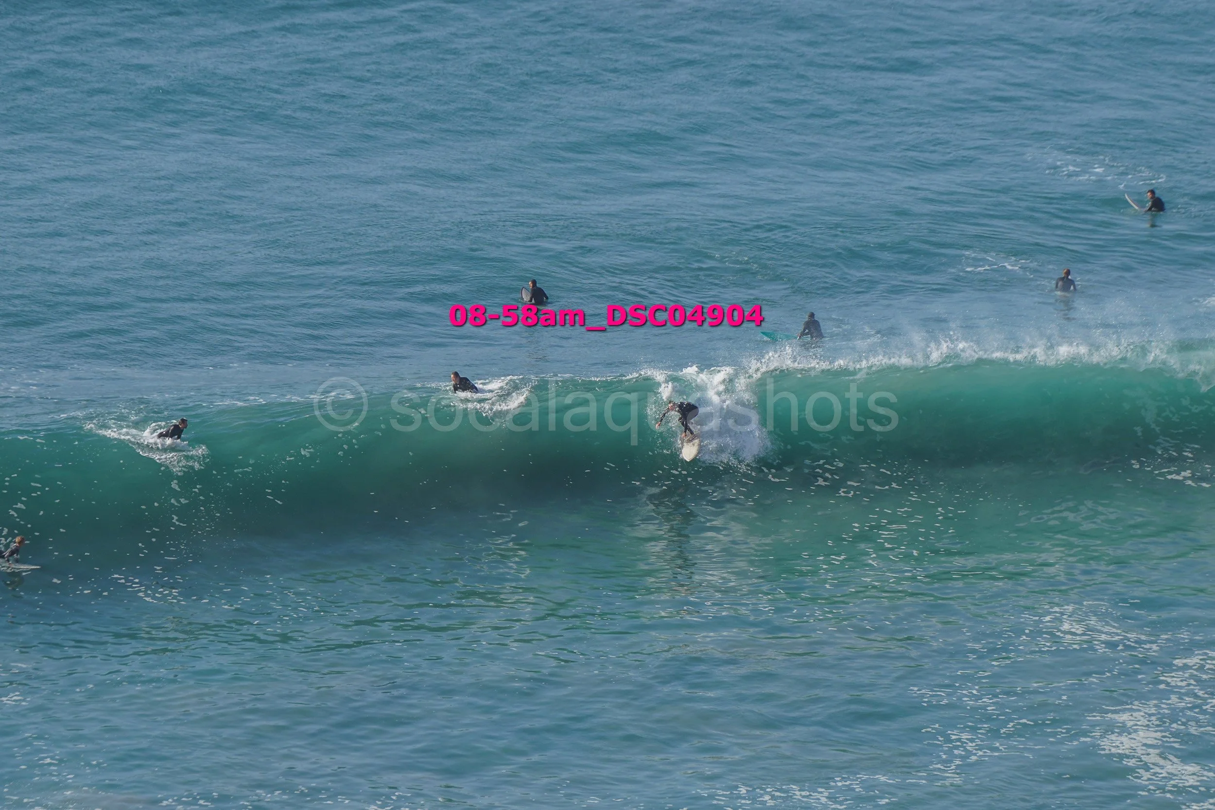 Multiple surfers in wetsuits riding and waiting for waves at the beach on a clear day.