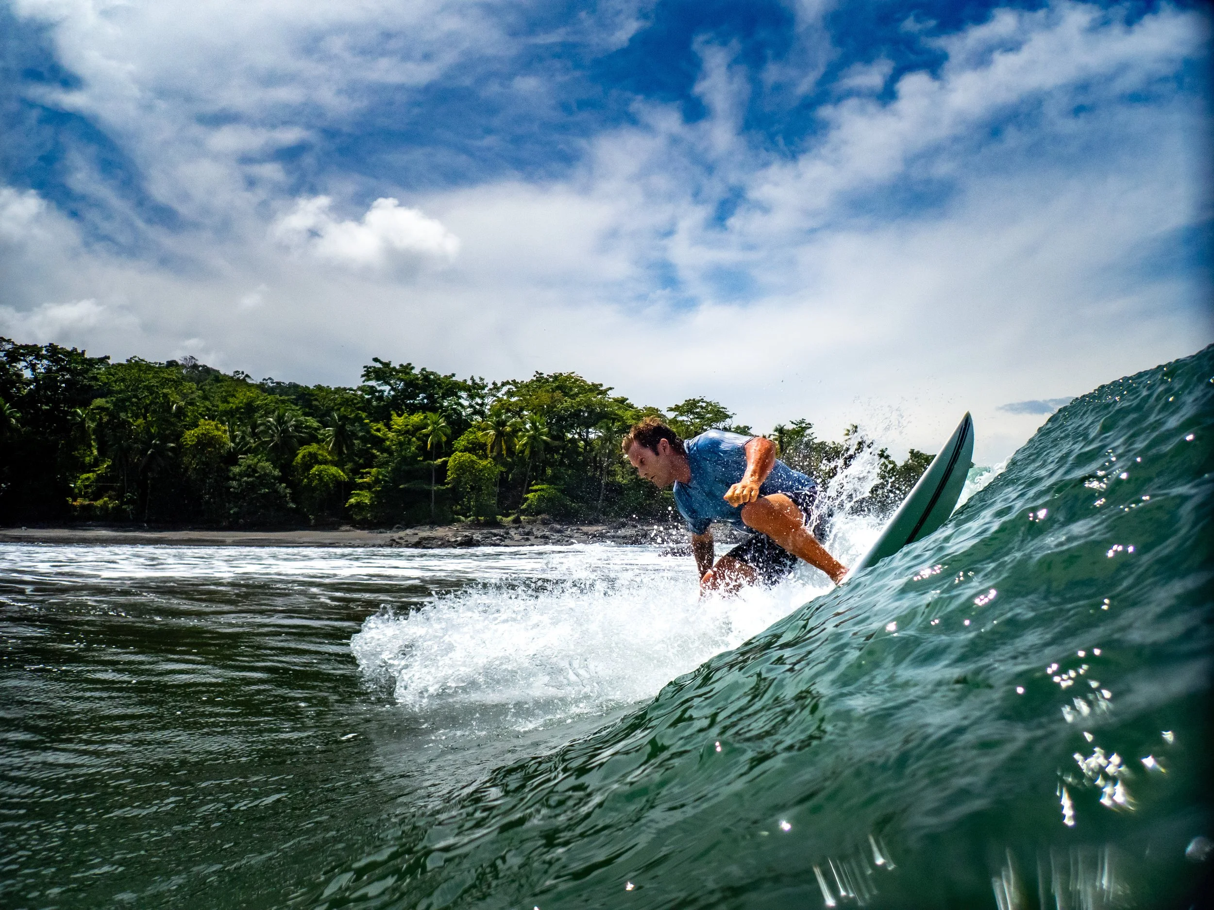 Man surfing on a wave near a lush tropical coastline under a blue sky with clouds.