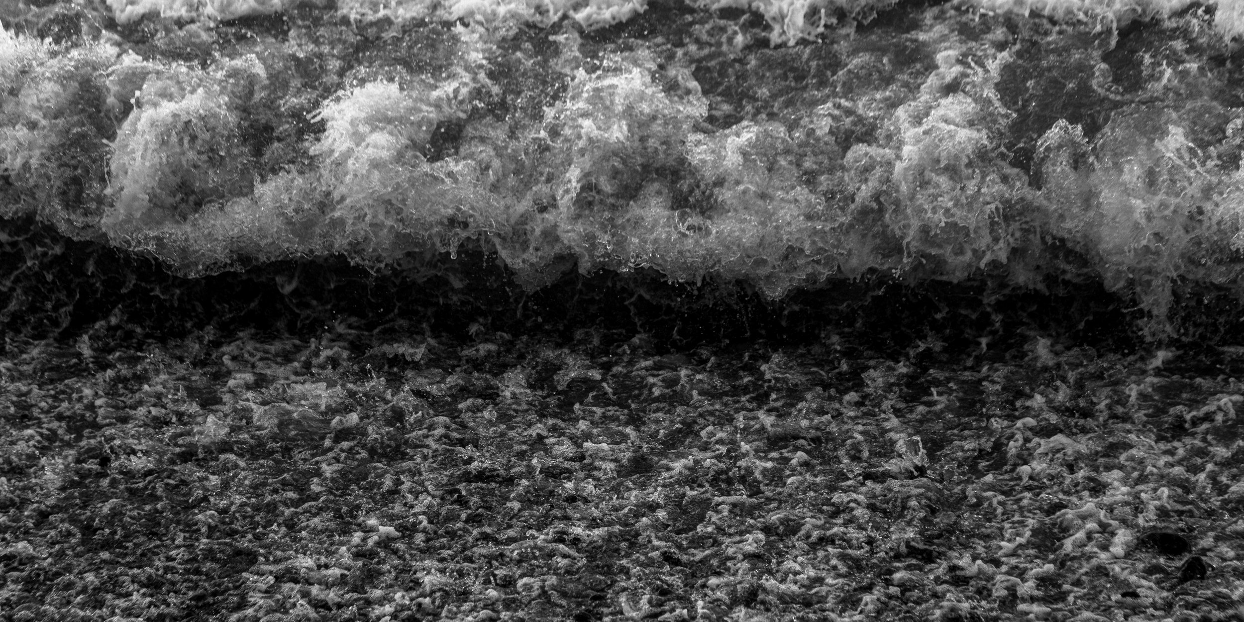 Black and white photo of ocean waves crashing onto a pebbled beach.