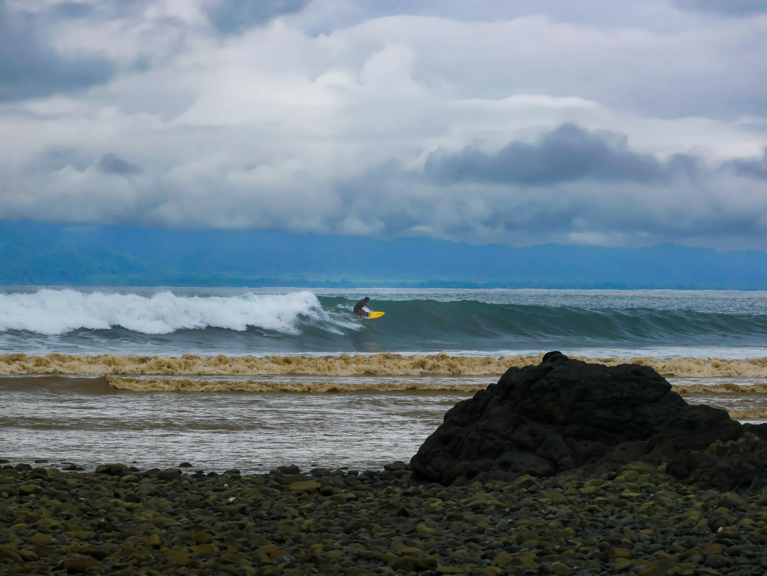 Surfer riding a wave on a cloudy day with rocky beach in foreground.