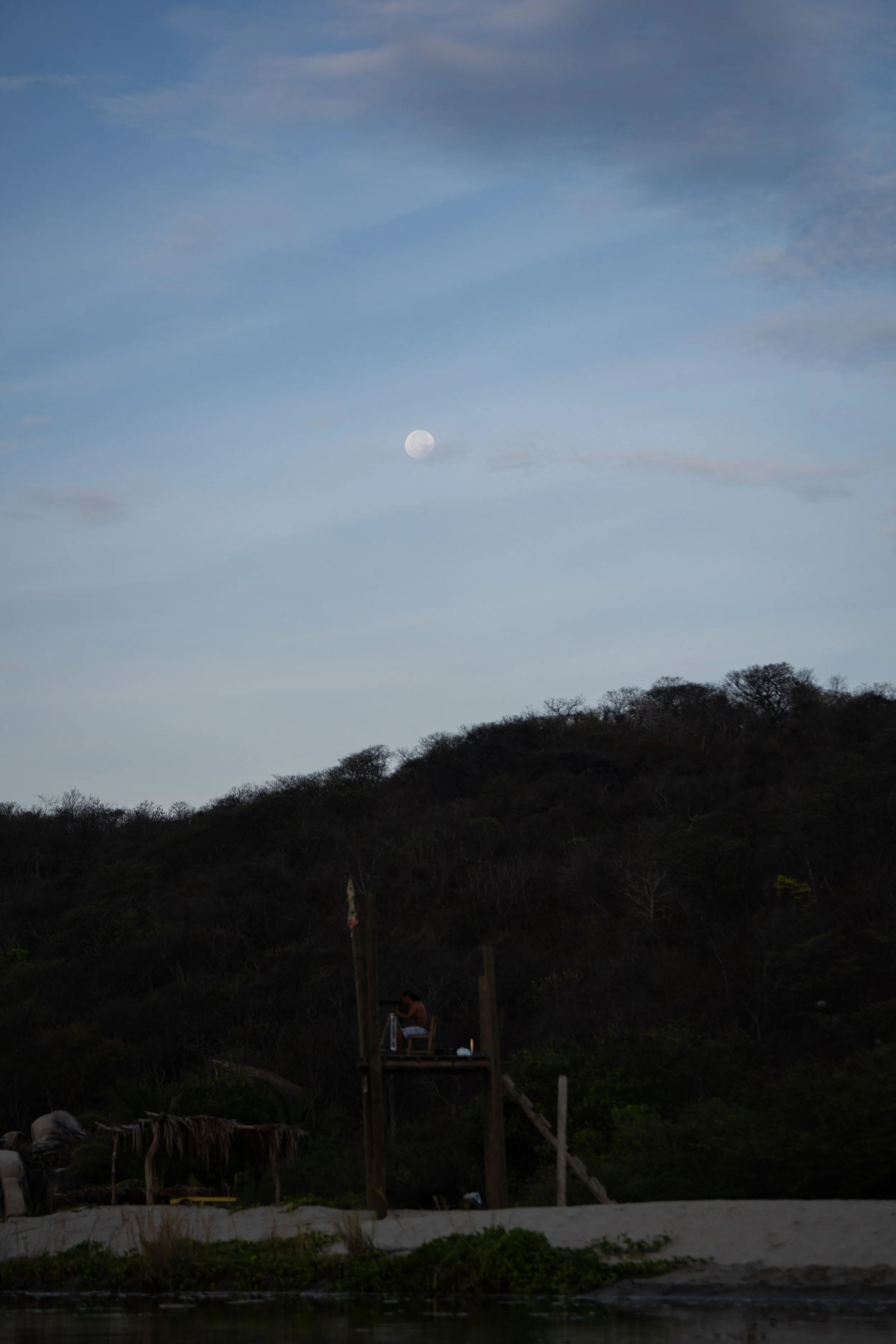 A landscape scene with a clear blue sky, the moon visible, and a hillside covered with trees. There is a small structure with a person sitting on a chair underneath, and the foreground shows part of a river or pond with vegetation.