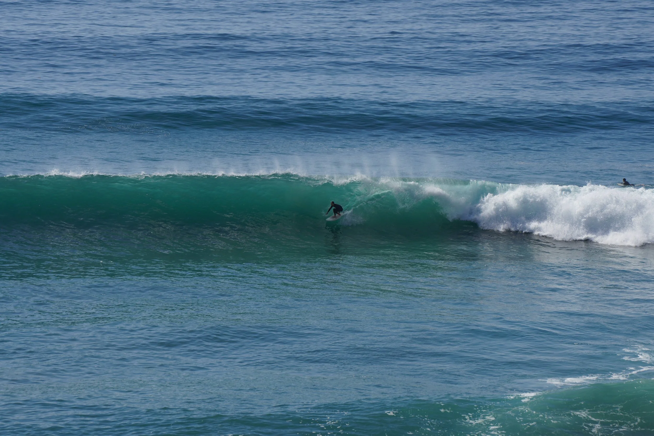 A person surfing on a green wave in the ocean, with another surfer visible in the distance.