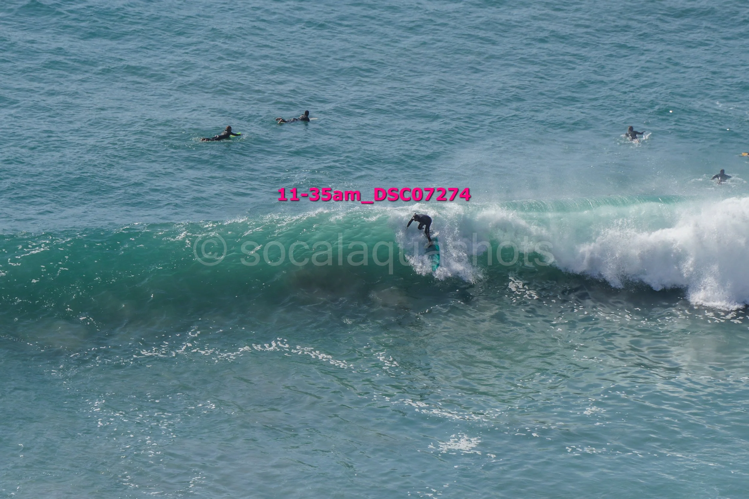 Person riding a surfboard on a wave with several people swimming in the water nearby.