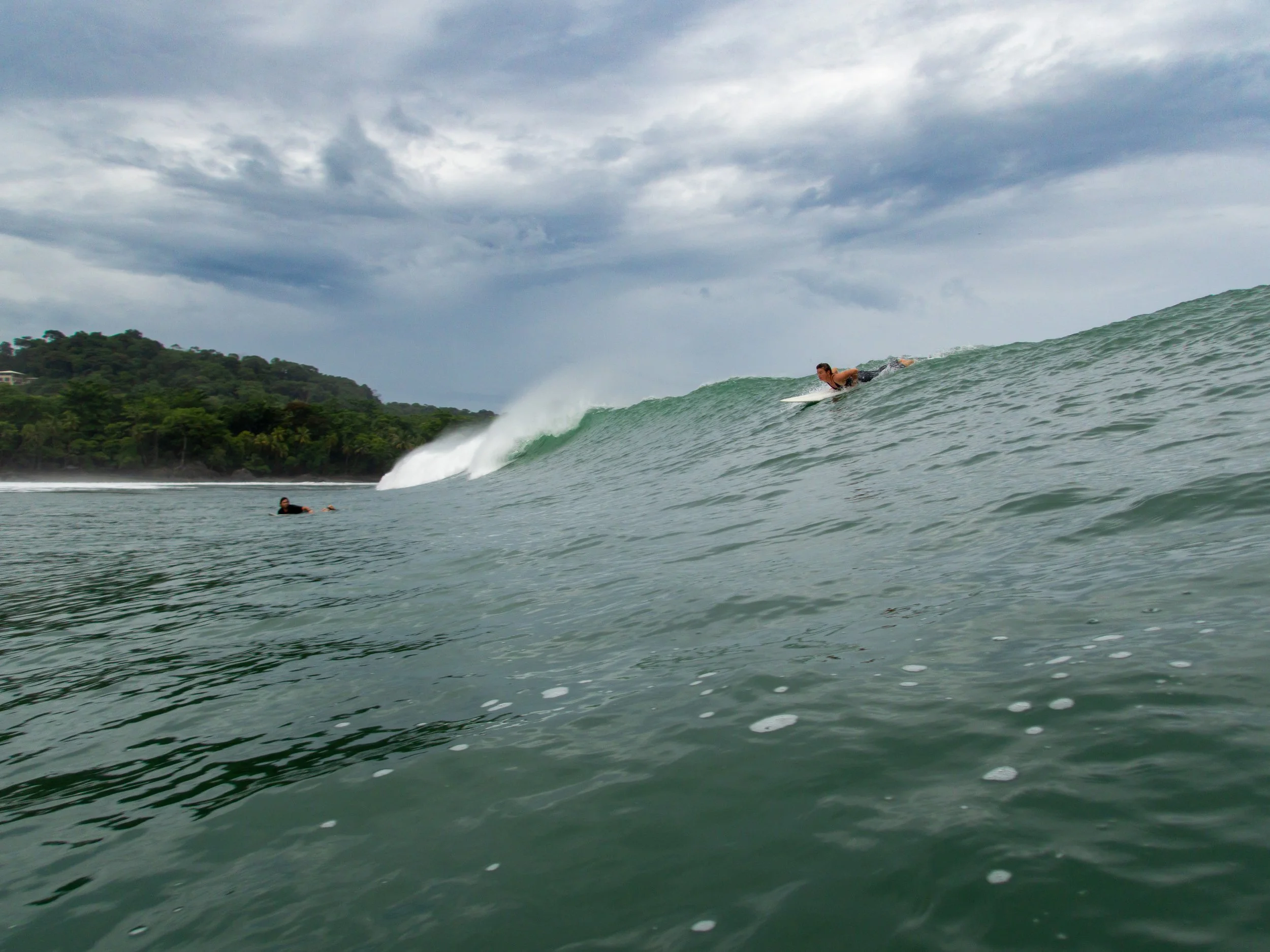 Surfer paddling on a wave with another person in the water, cloudy sky, and distant shoreline in the background.