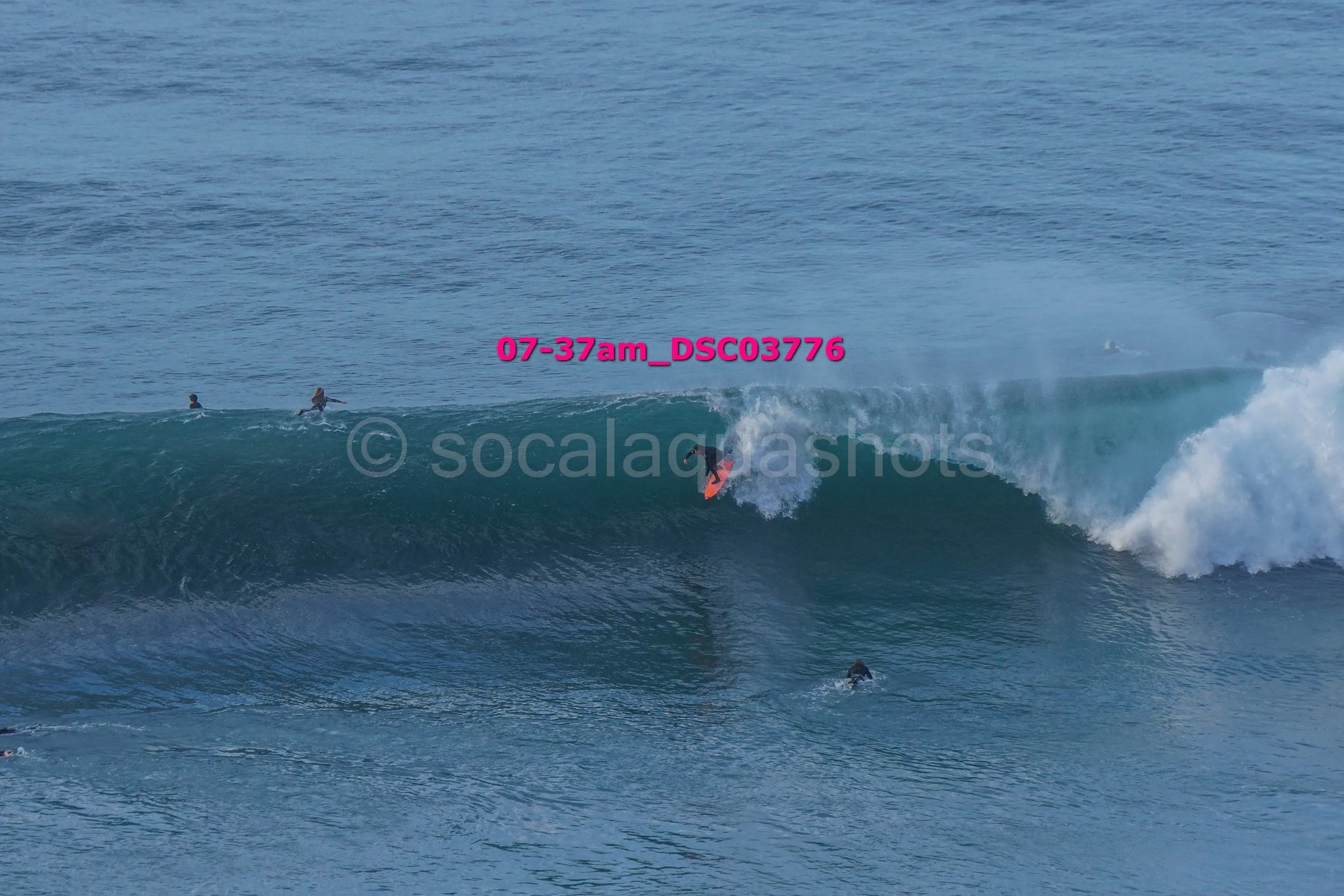 A person surfing a large wave with other surfers in the water nearby on a clear day.