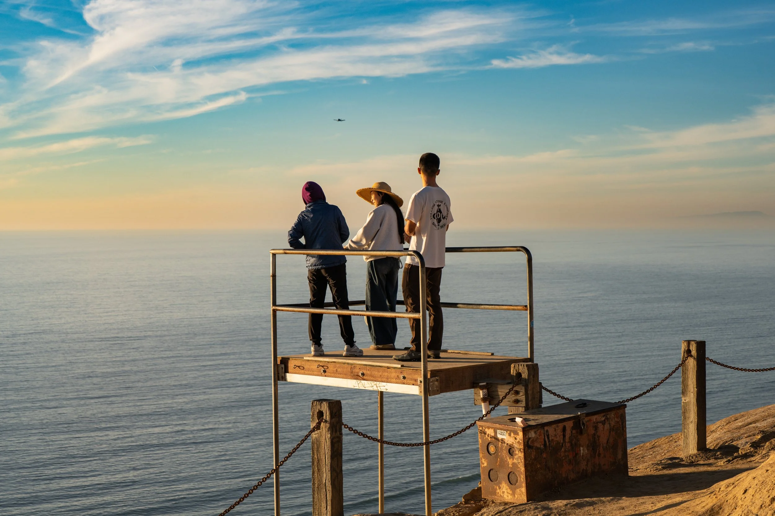 Three people standing on a small wooden dock with a metal railing beside a calm ocean during sunset, with an airplane flying in the sky above.