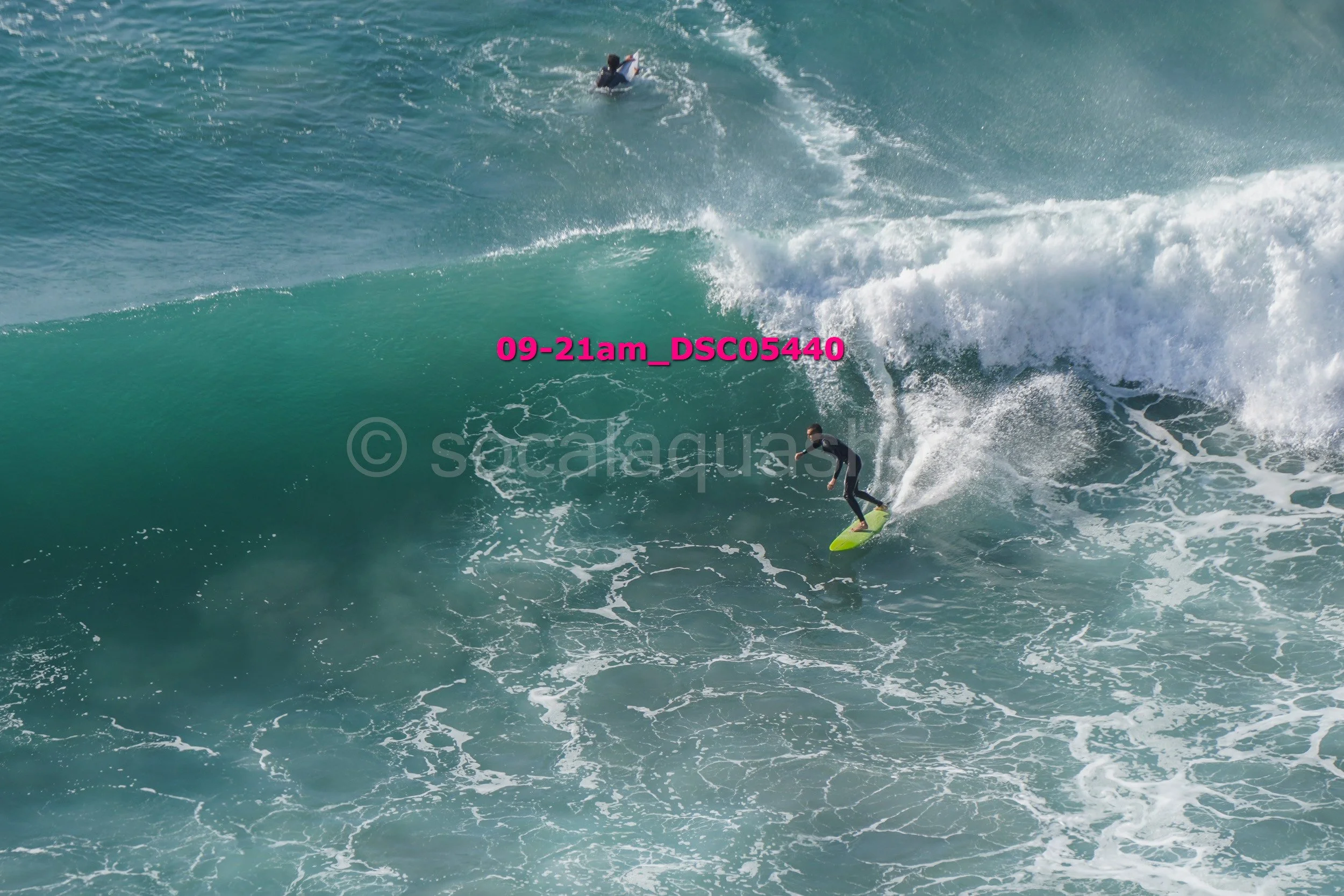 A person surfing on a large wave in the ocean with another surfer visible in the background.