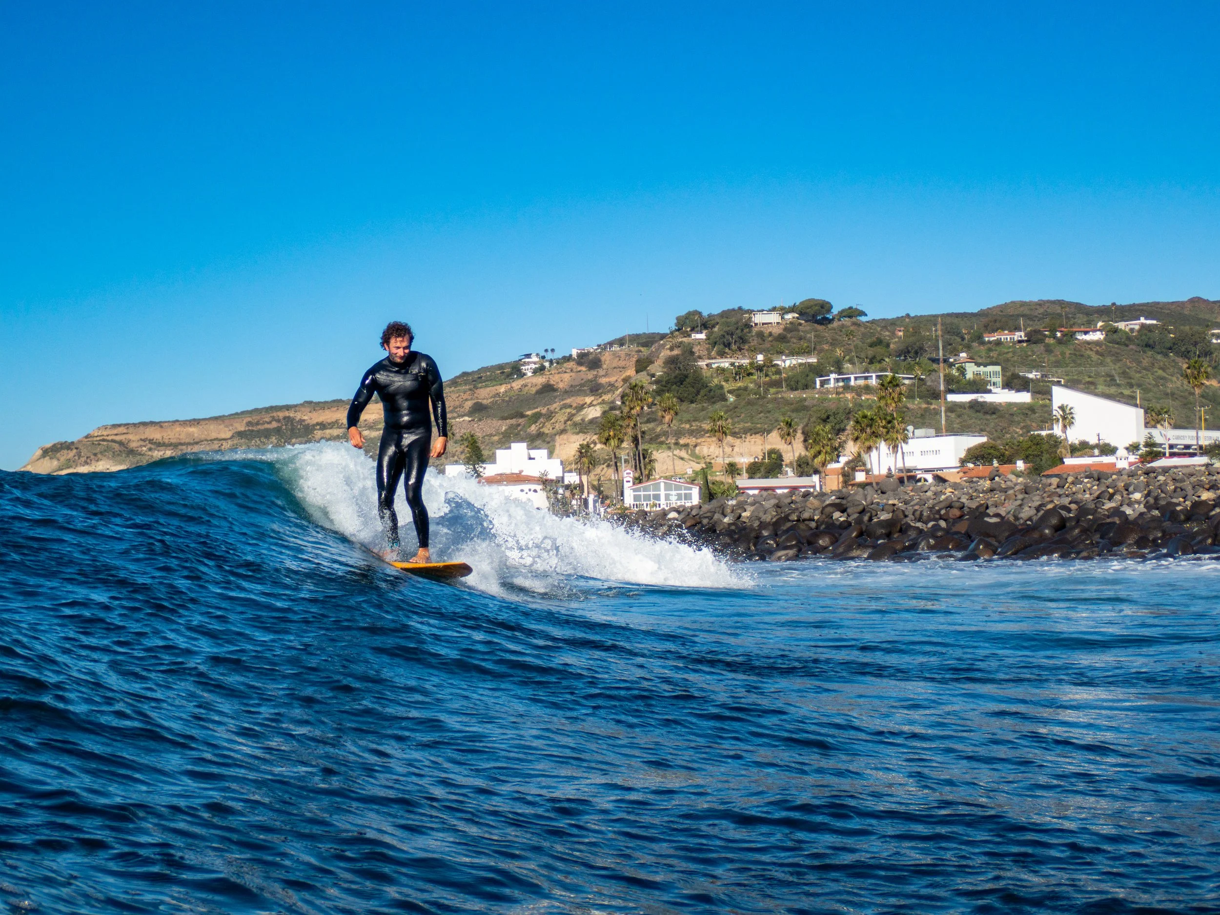 A person in a black wetsuit surfing on a wave near the shoreline with houses and hills in the background.